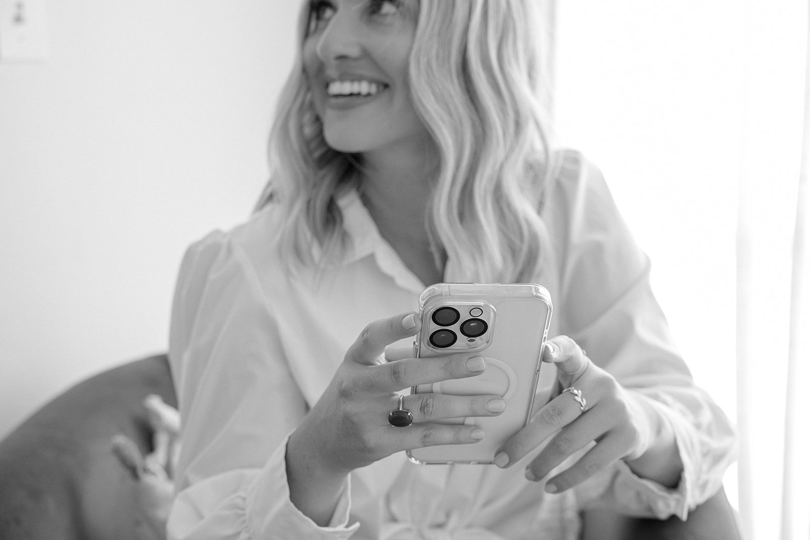 A woman with wavy blonde hair smiling while looking to her right, holding a smartphone in her left hand, with a bright background.