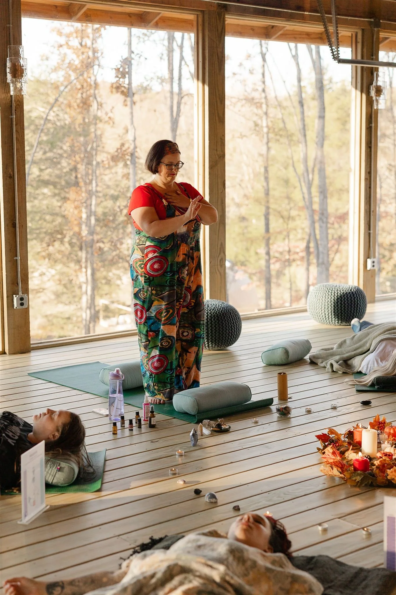 A woman in colorful pants and a red top leads a meditation session in a bright, wooden room with large windows showing trees outside. She places her hands on her chest with eyes closed, while people lie on mats with their eyes closed, surrounded by candles, plants, and meditation props.