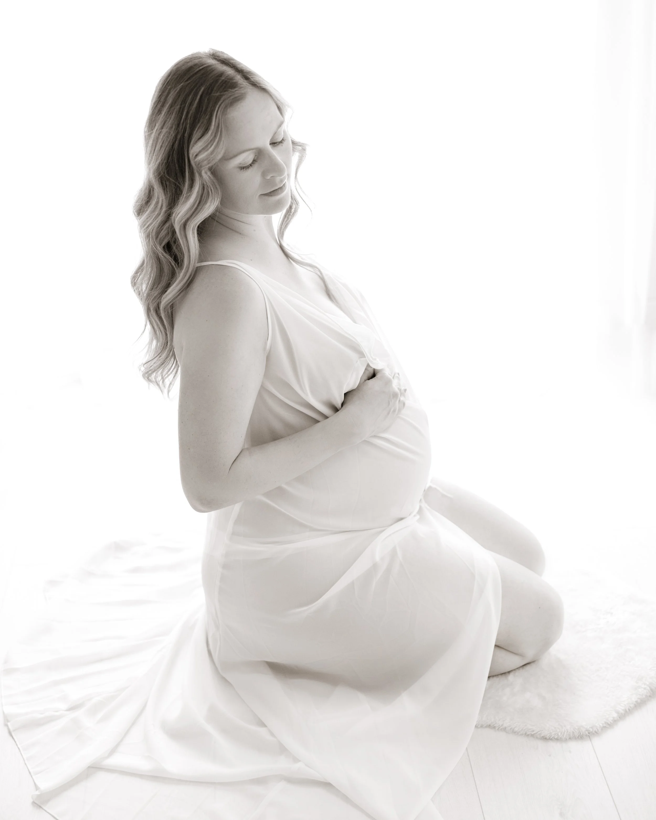 A pregnant woman with long wavy hair kneeling on a soft rug, wearing a flowing light-colored dress, with her hands gently resting on her belly, in a bright, softly lit room.