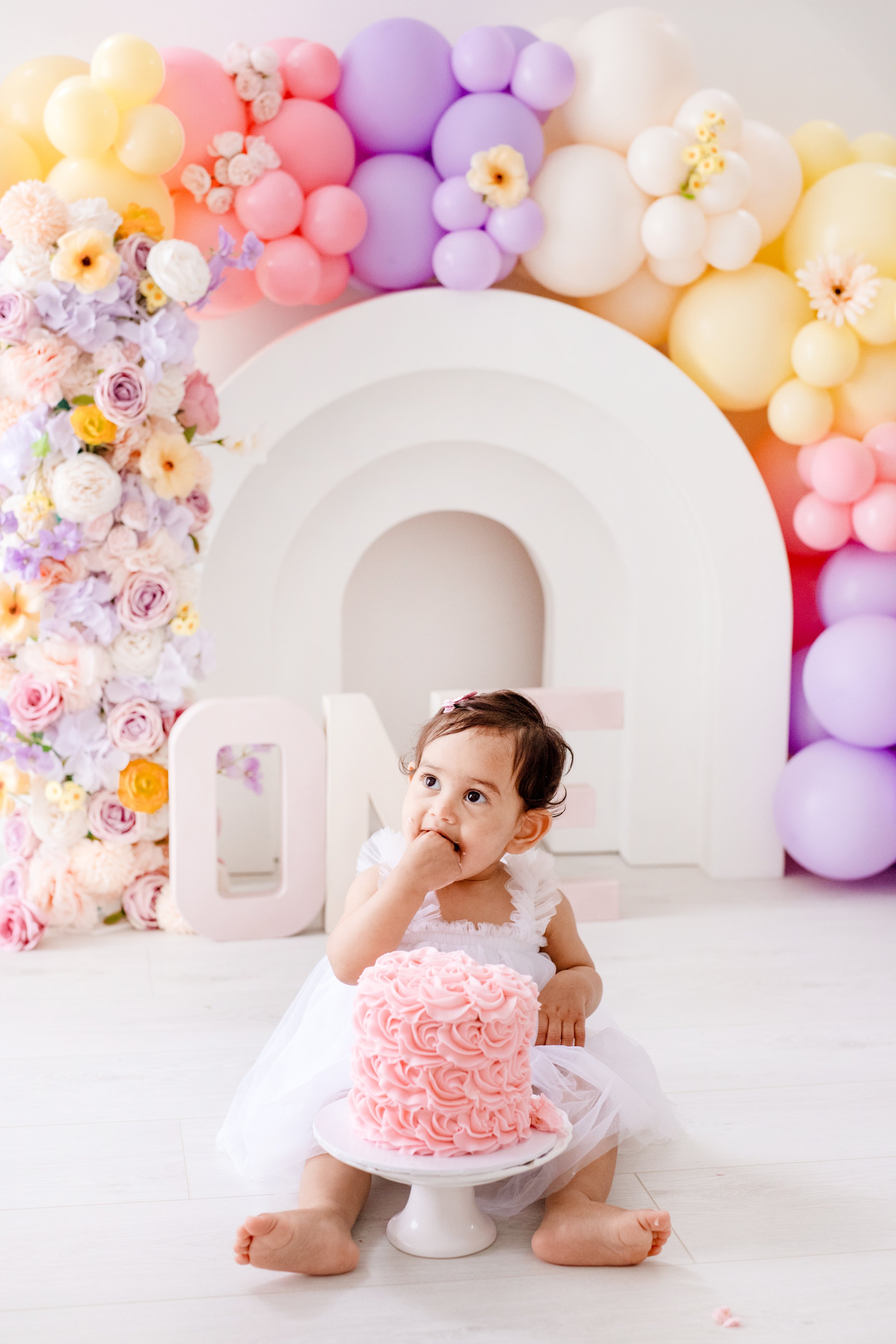 A young girl in a white dress sitting on the floor, eating pink frosted cake with rosette design, with a birthday cake in front of her. .  first birthday cake smash Mississauga