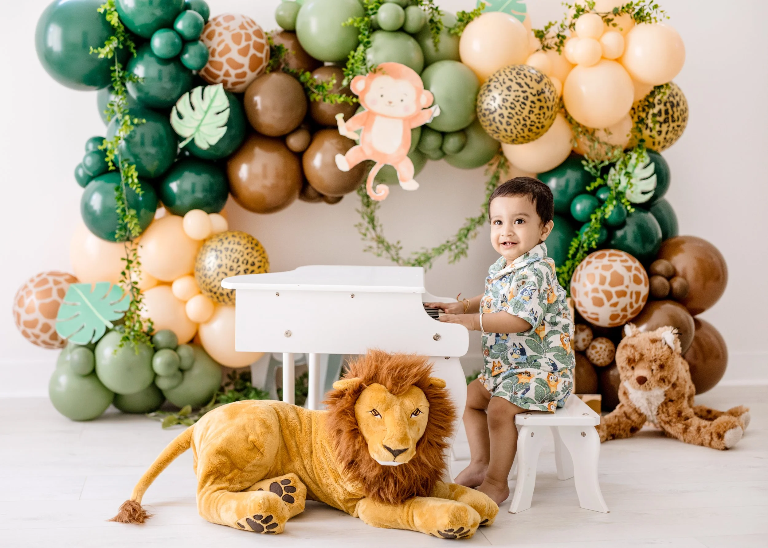 A young boy playing piano surrounded by plush lion and leopard toys, with a colorful jungle-themed balloon backdrop.  first birthday cake smash Mississauga