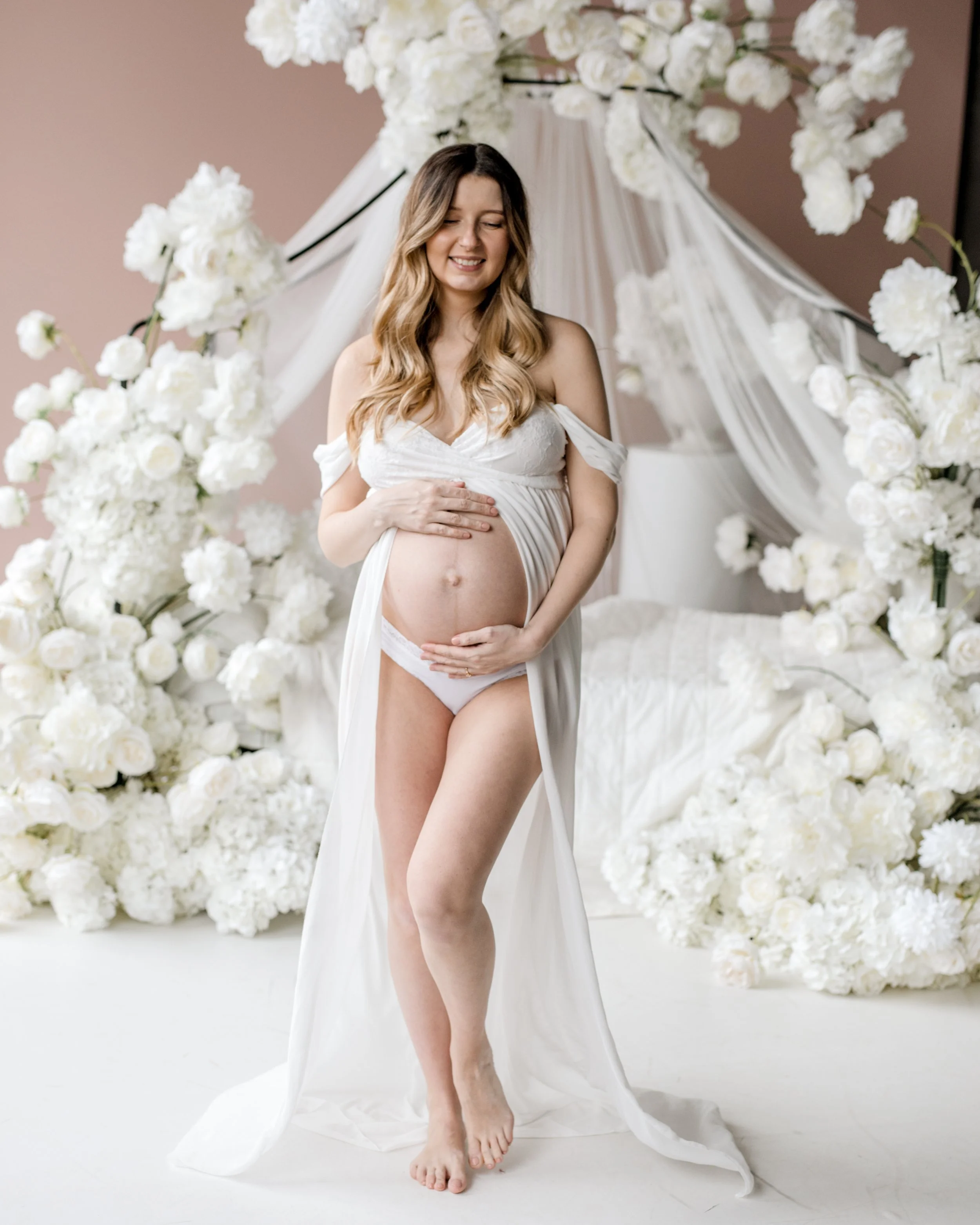 Pregnant woman in a white maternity gown standing in front of a floral backdrop, smiling and holding her belly – professional maternity photography in Toronto Mint Room Studios.