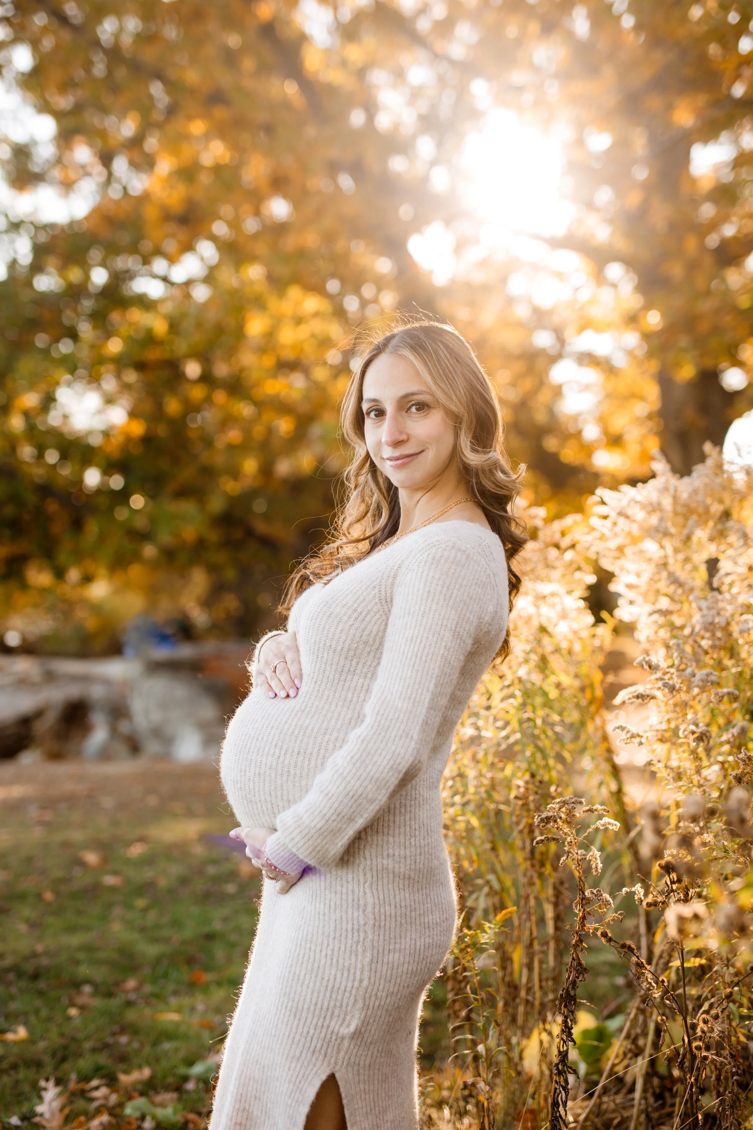 A pregnant woman standing outdoors during fall, with trees and golden sunlight in the background, wearing a beige knitted dress.