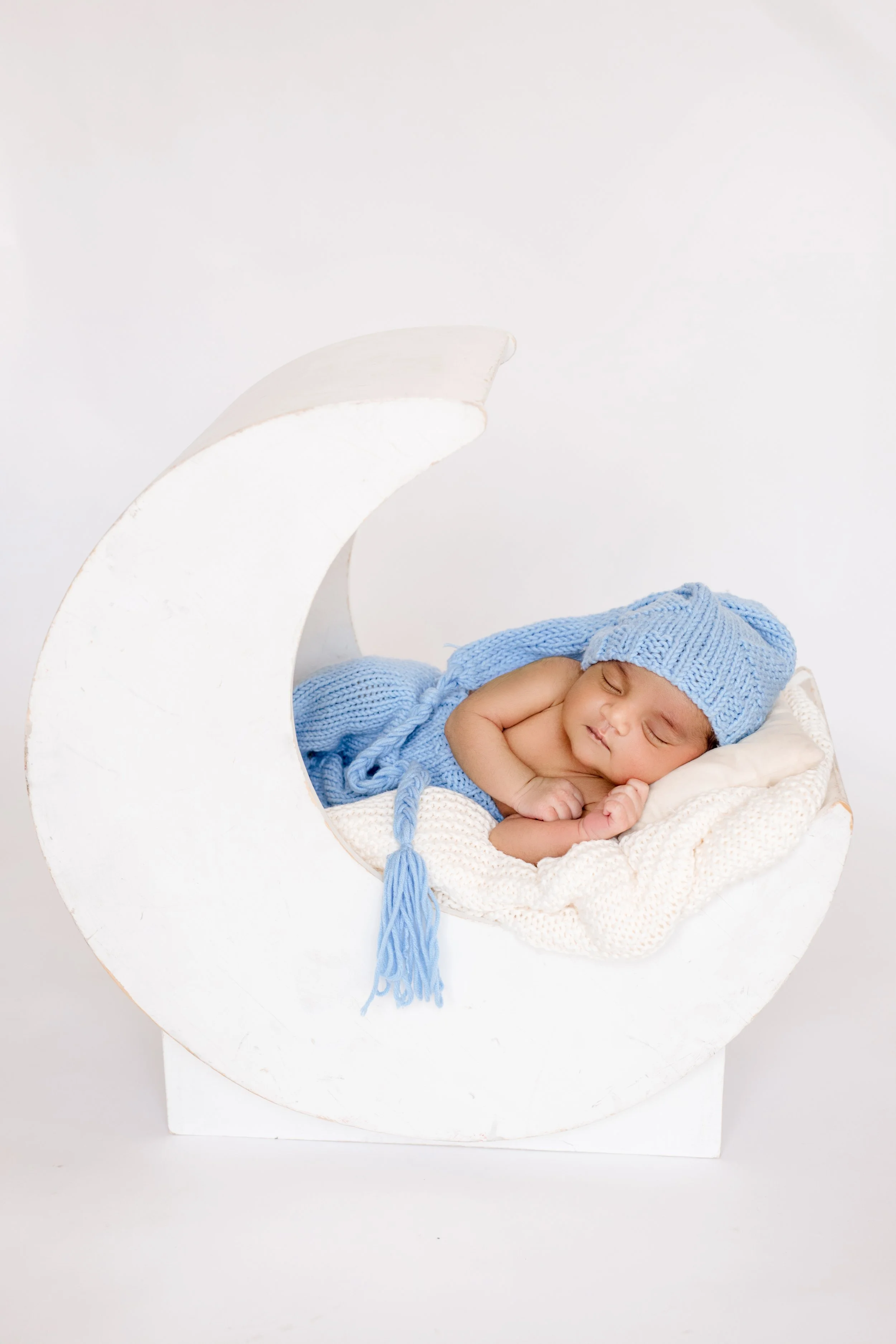 A sleeping baby with a blue knit hat and matching blanket, lying on a white blanket inside a white crescent moon prop.