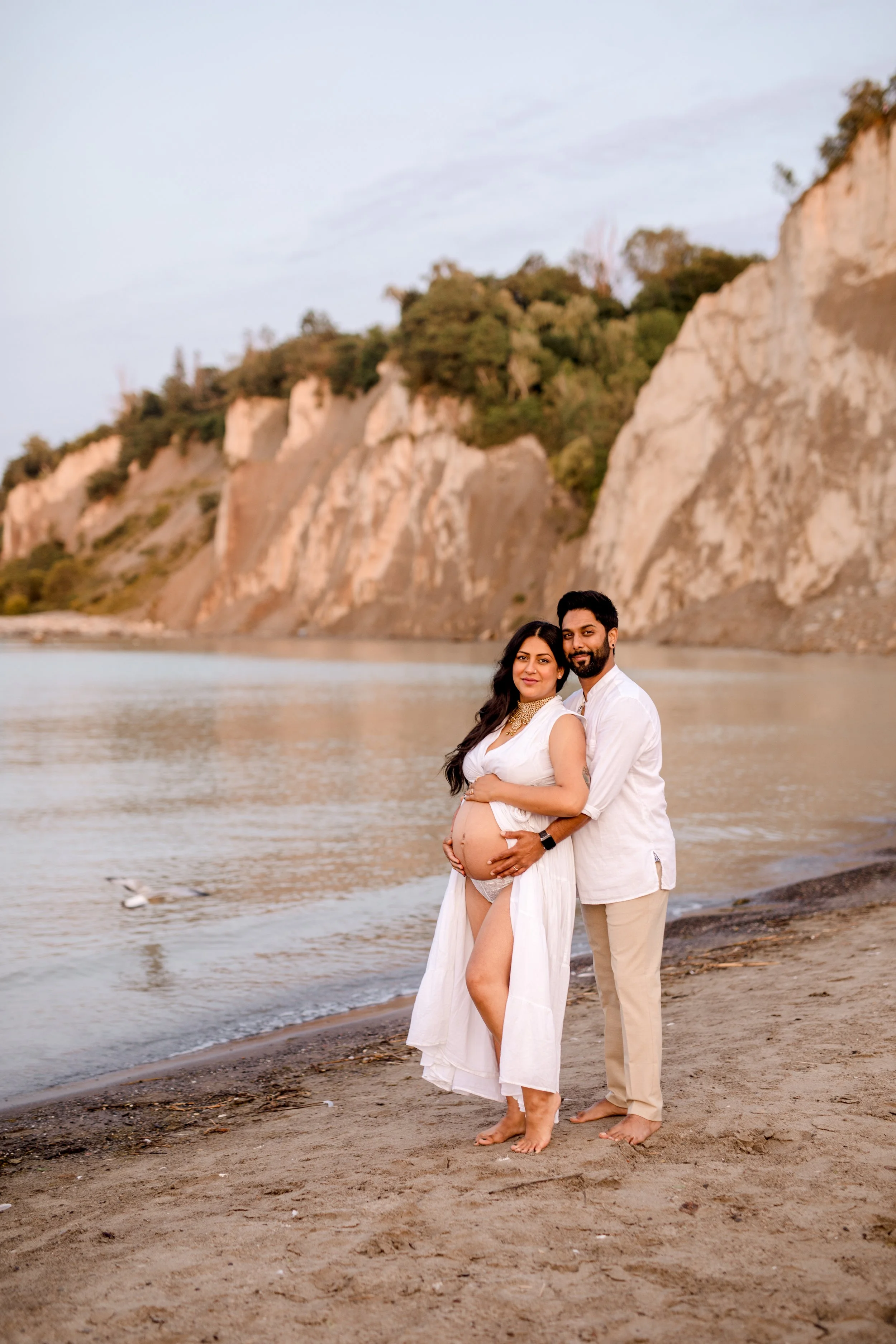 Pregnant woman in a white dress and her partner in beige pants and white shirt standing on a beach at Scarborough Bluffs, woman cradling her belly and man holding her waist, both smiling – professional maternity photography in Toronto.