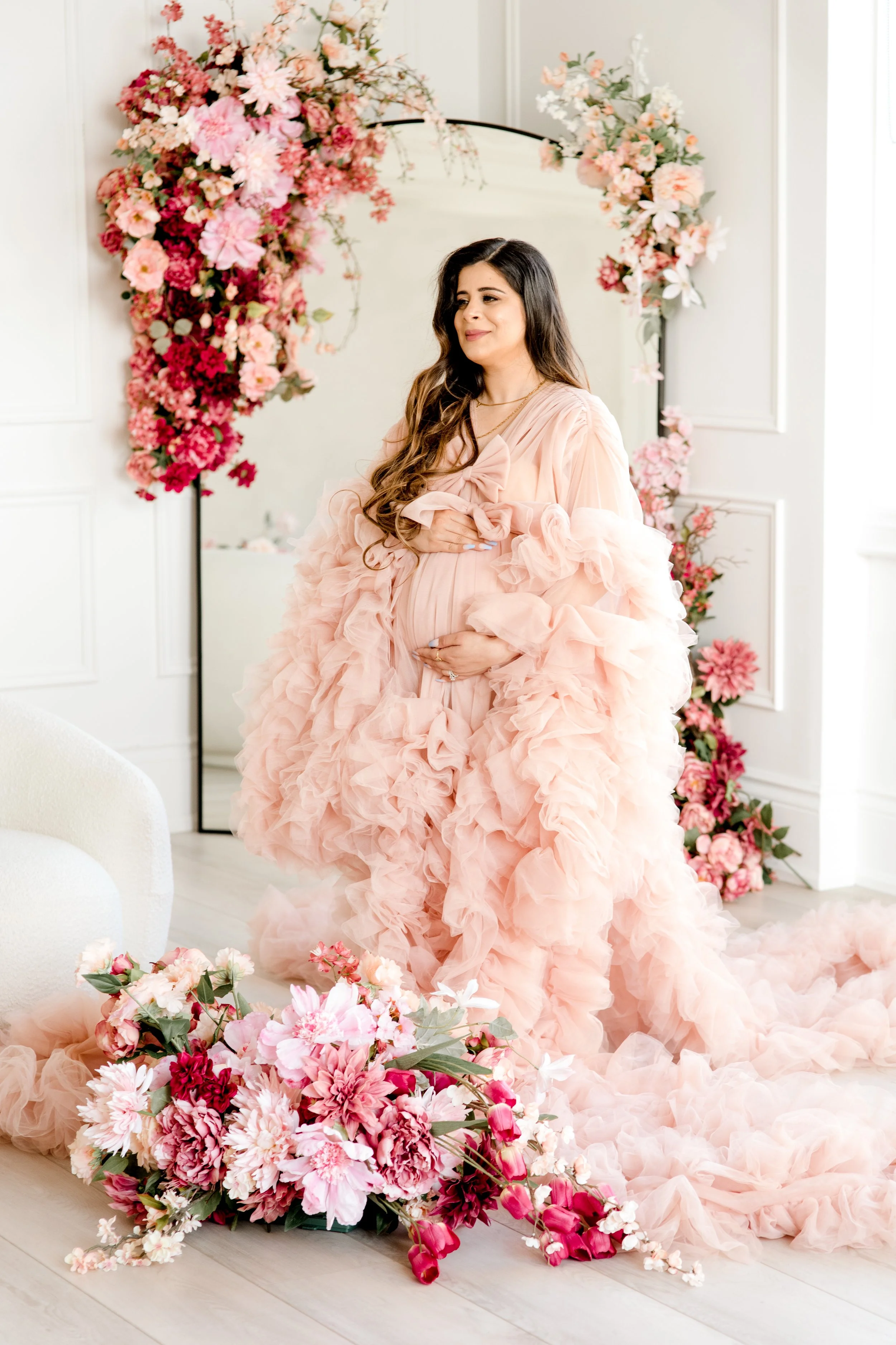 Pregnant woman in a flowing pink ruffled gown in a decorated floral studio.