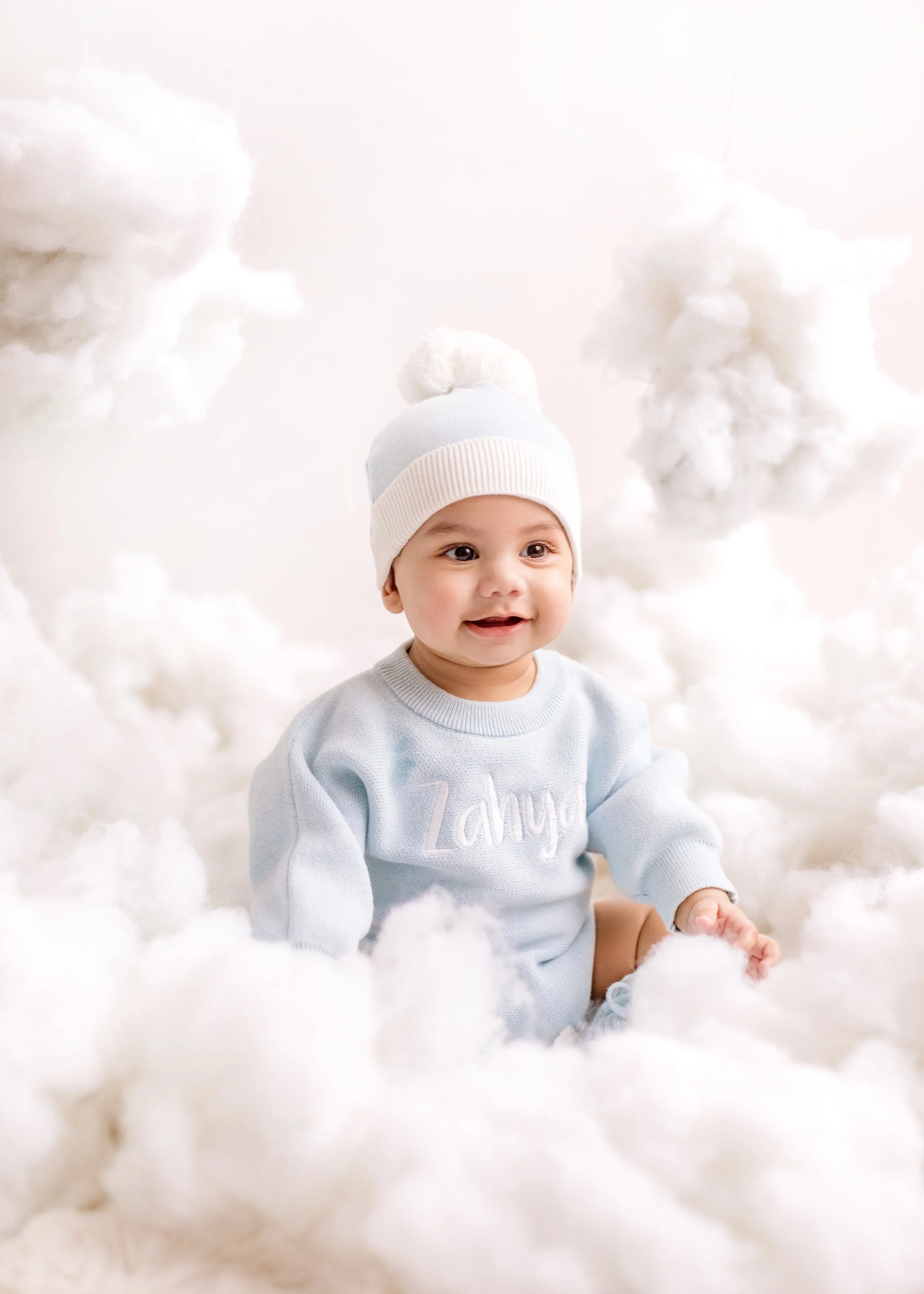 A smiling baby wearing a white knit hat and a light blue sweater with the name 'Zahya' embroidered on it, sitting among fluffy white clouds in a studio setting. Six month milestone shoot Mississauga