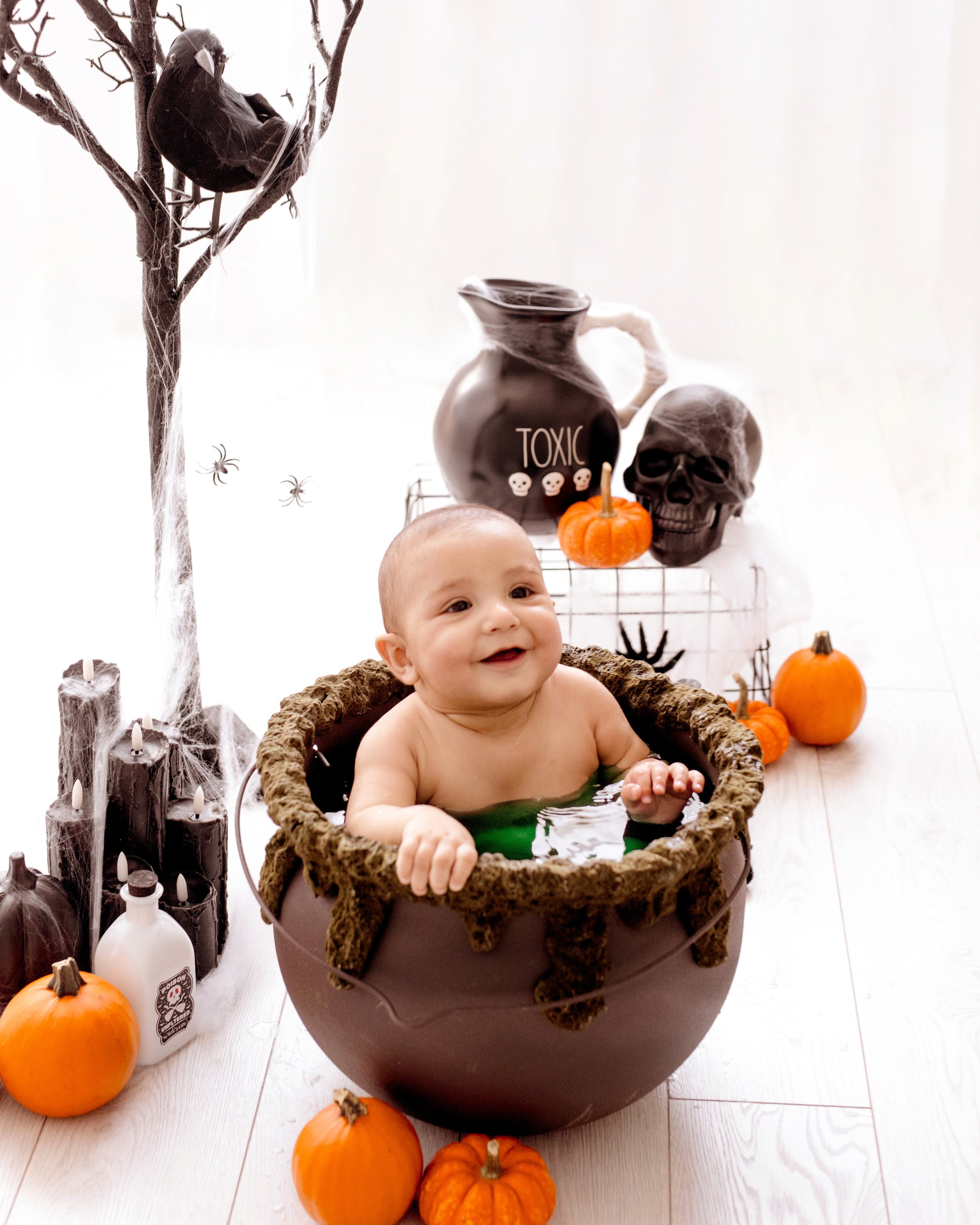 Baby sitting in a Halloween-themed cauldron with pumpkins and spooky decorations in the background. professional baby photography in Mississauga.