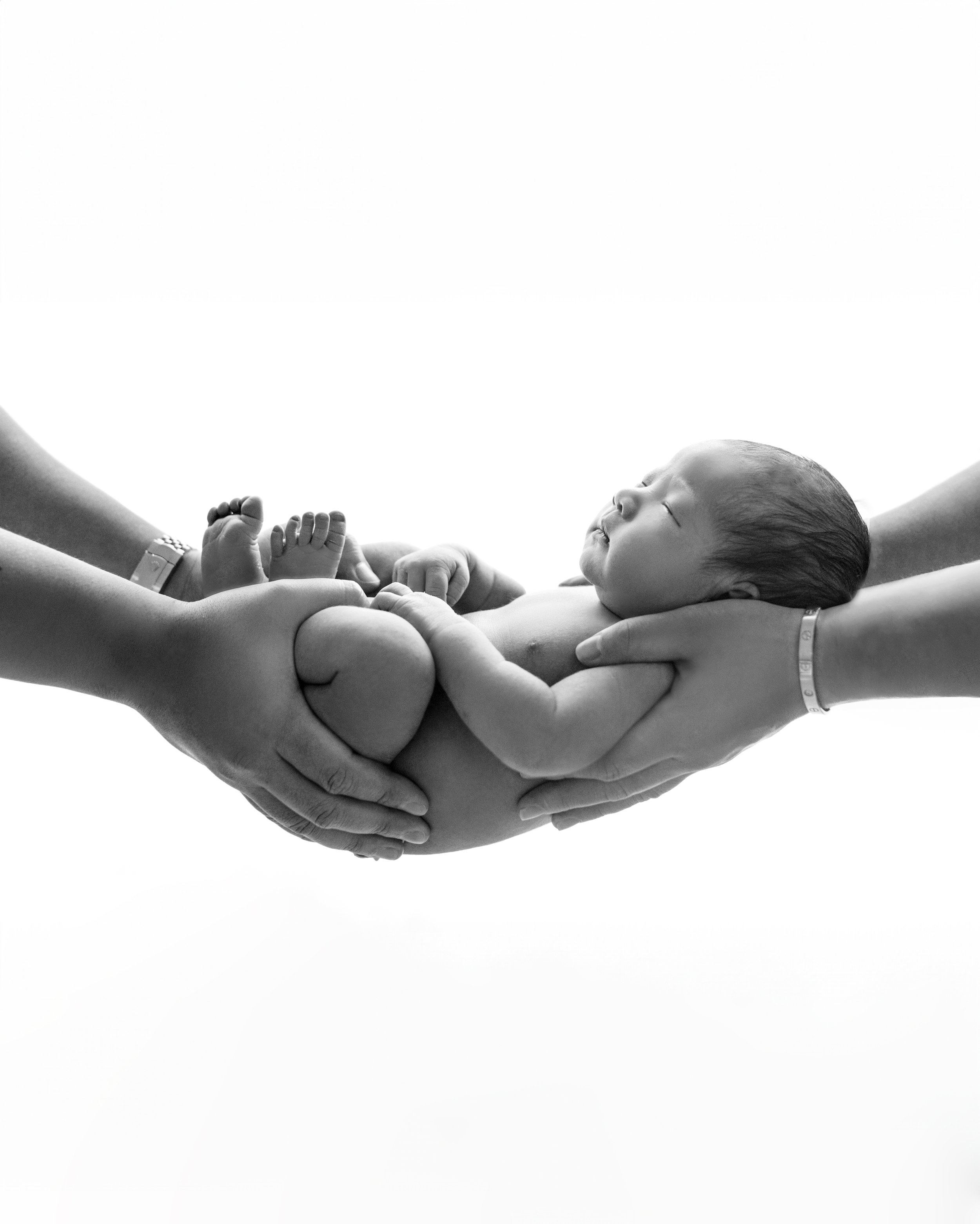 Black and white photo of a newborn baby cradled in multiple hands, with the baby's head resting on one hand and feet being gently held by others.
