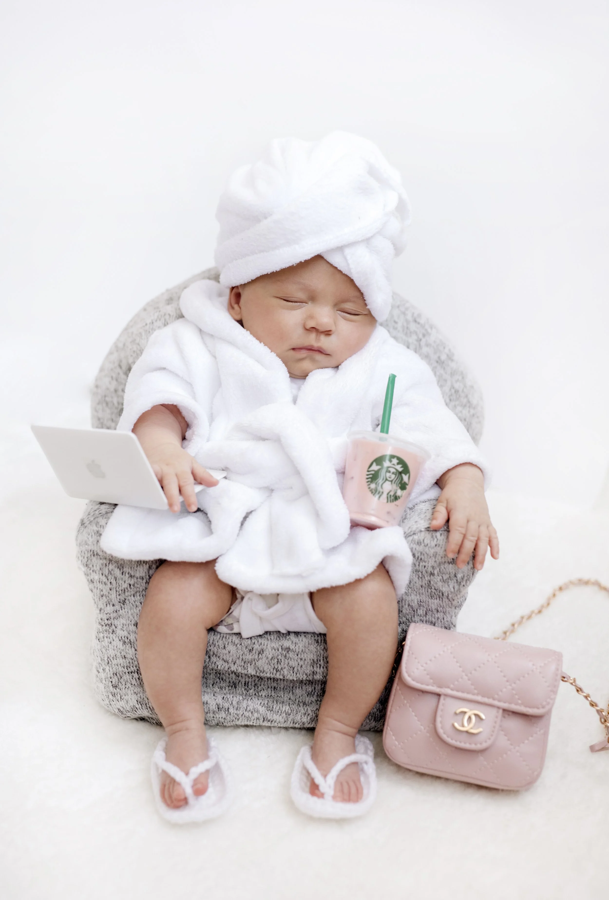 Sleeping newborn baby in a white towel robe and head wrap, sitting in a gray chair, holding a pink Starbucks drink and a MacBook, with white sandals and a pink Chanel quilted purse nearby – professional newborn photography in Mississauga.