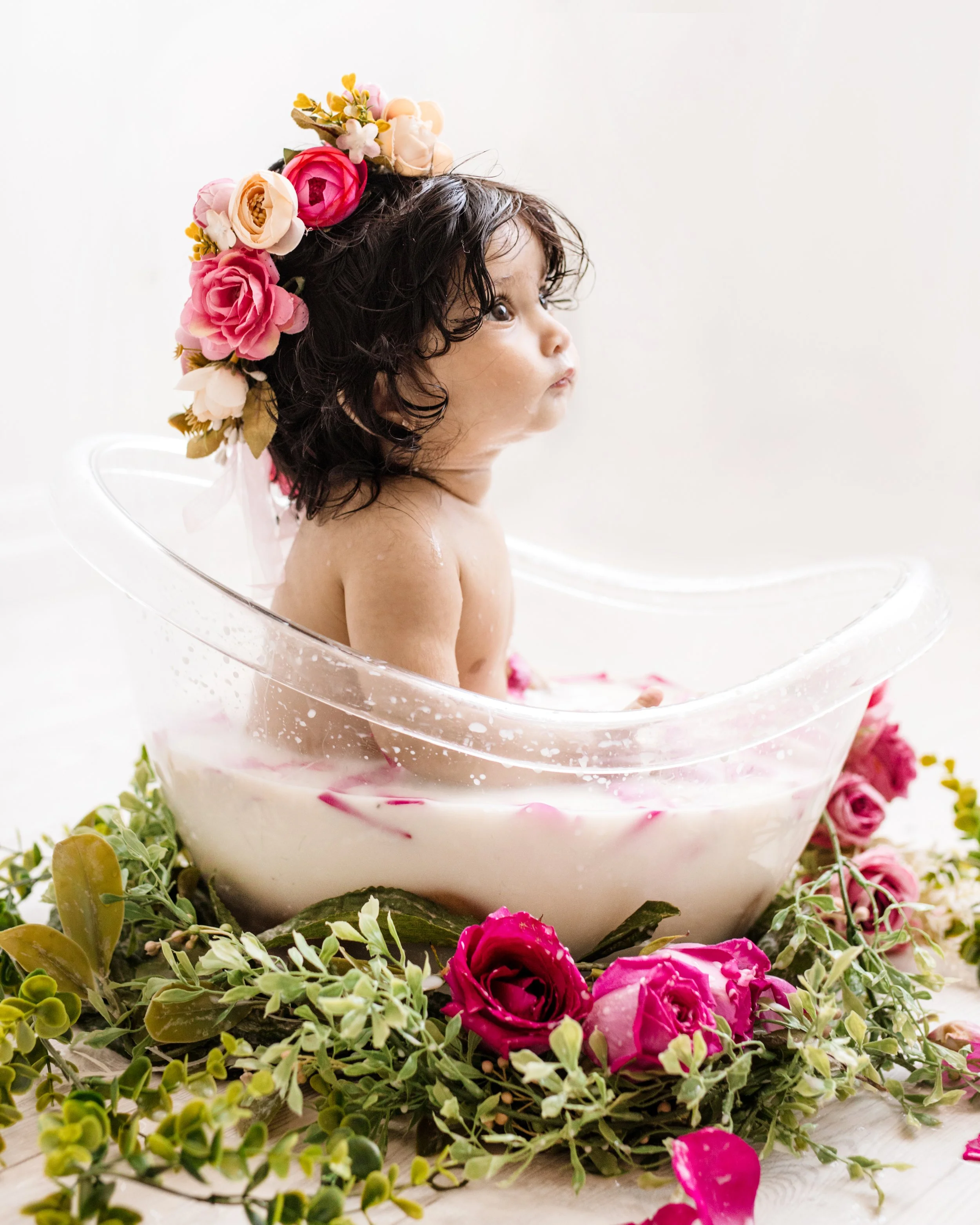 Six-month-old baby sitting in a clear glass bowl filled with water, wearing a floral crown, surrounded by pink and white flowers and green foliage – professional baby photography in Mississauga.
