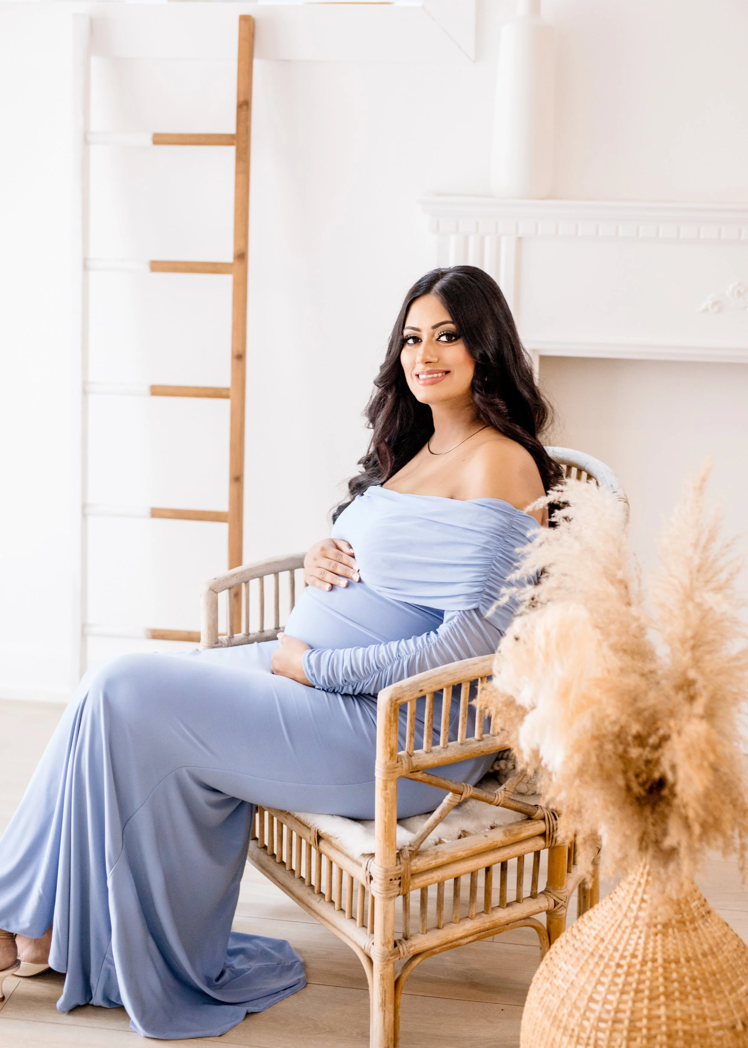 A pregnant woman sitting on a wicker chair in a bright, minimalistic room, wearing a long, elegant, off-the-shoulder blue dress, with her hand resting on her belly, smiling at the camera. There is a decorative vase with pampas grass next to her.