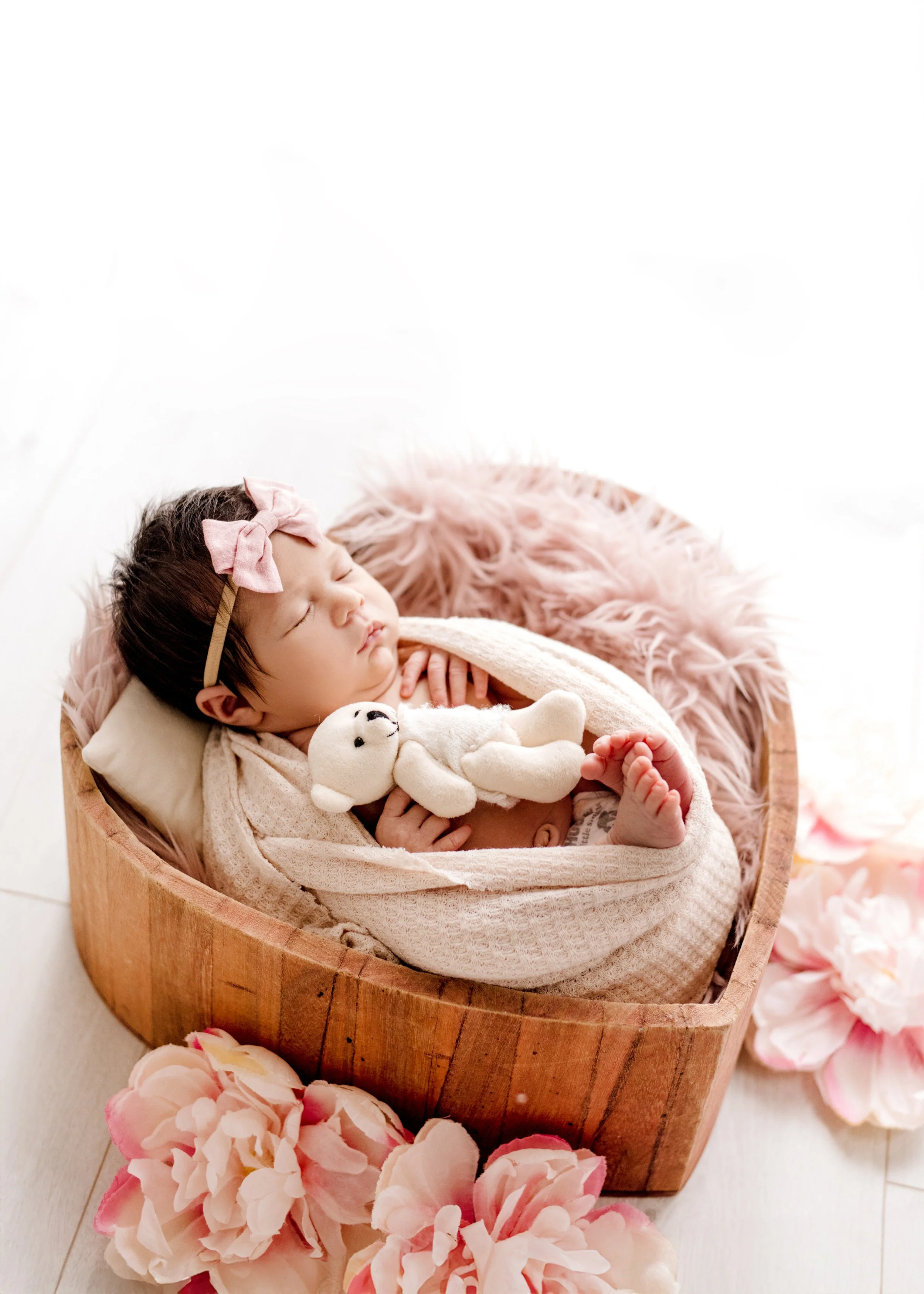Newborn baby girl sleeping in a wooden bowl, wrapped in a beige blanket, surrounded by pink peony flowers, holding a white stuffed bear – professional newborn photography in Mississauga studio.