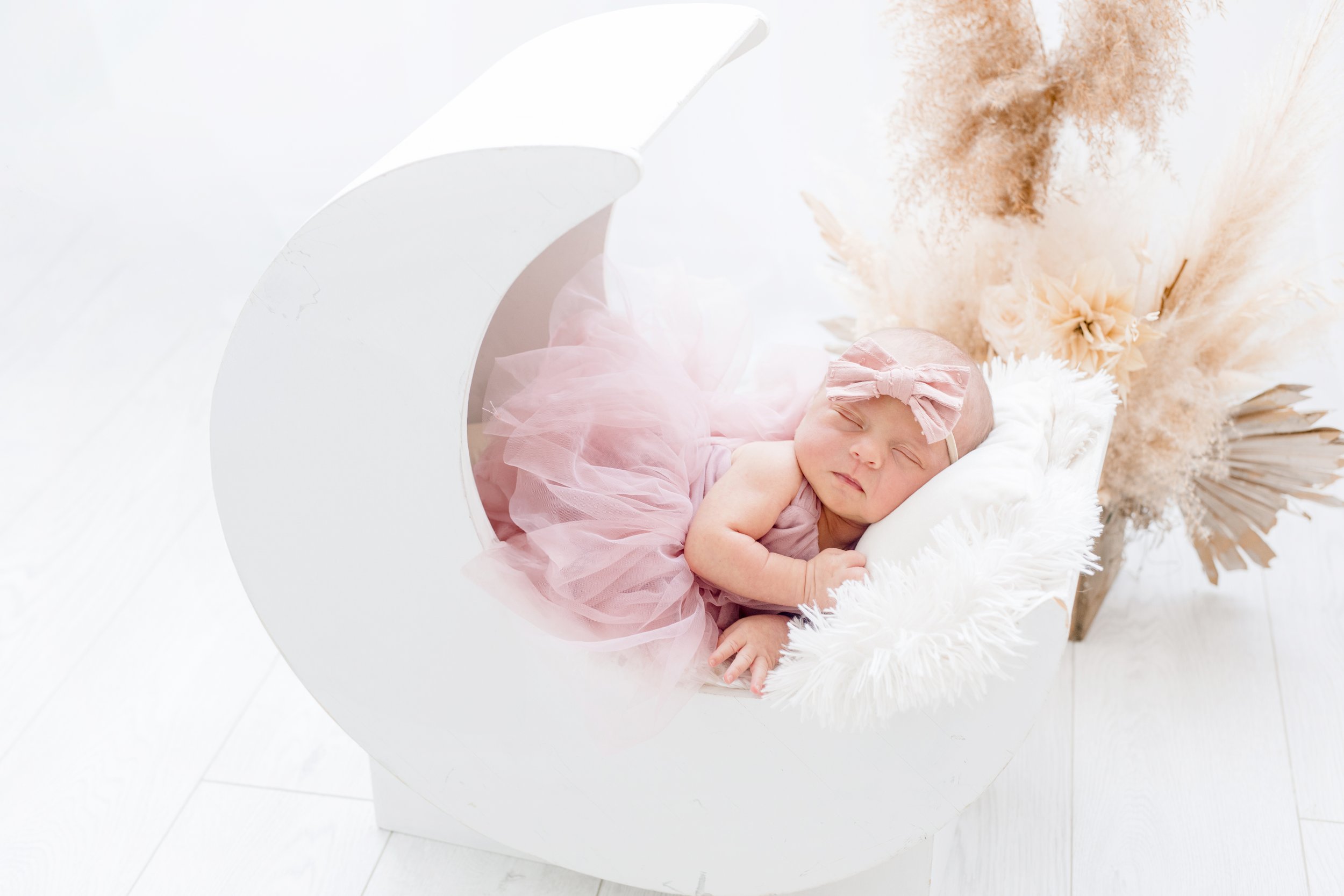Sleeping newborn baby girl with pink bow headband, lying on her side in a crescent moon-shaped white bed, surrounded by neutral pampas grass and dried flowers – professional newborn photography in Mississauga.