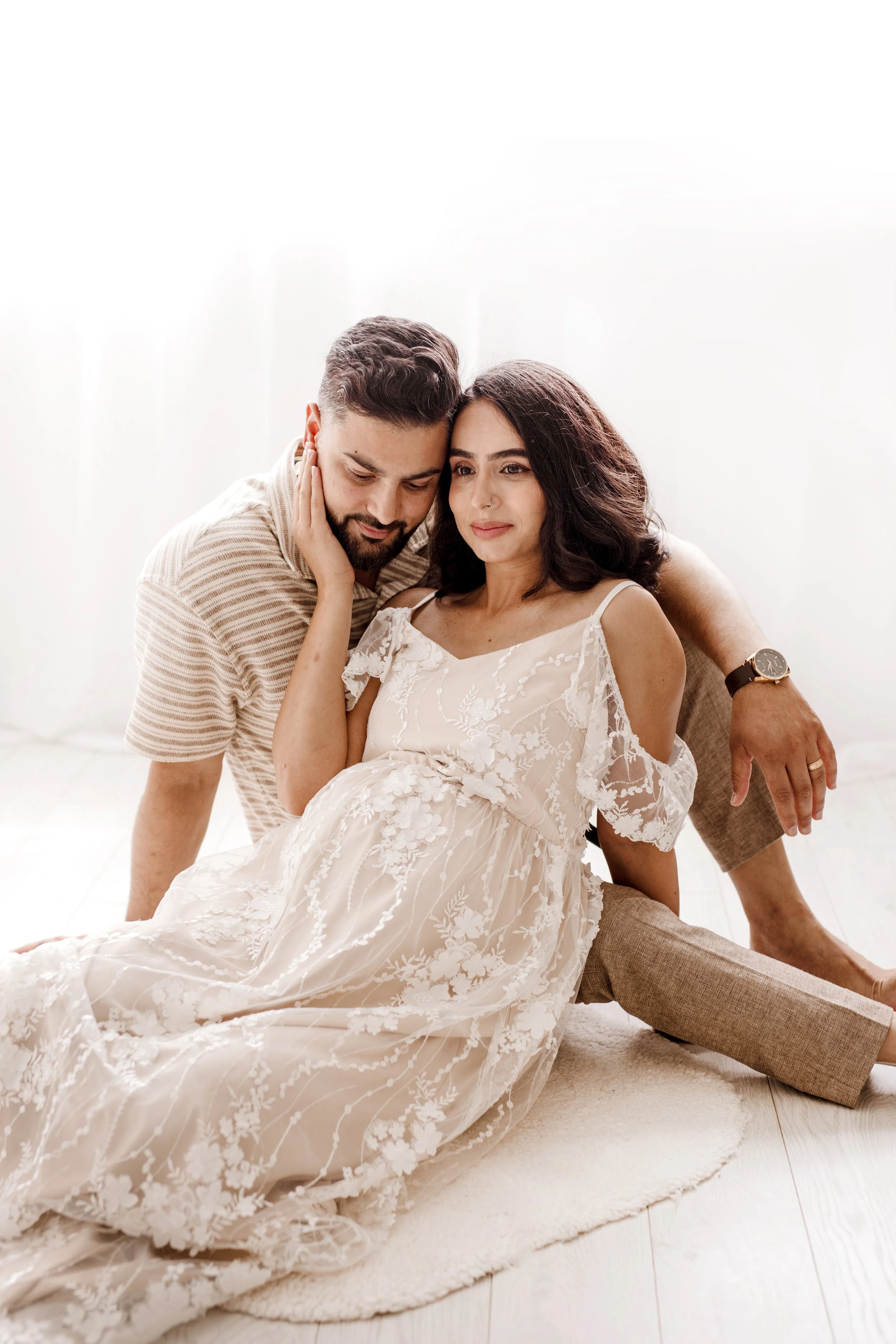 Pregnant couple sitting on a light-coloured rug indoors, woman in a white lace dress and man in a striped shirt and beige pants, embracing each other in bright natural light – professional maternity photography in Mississauga.