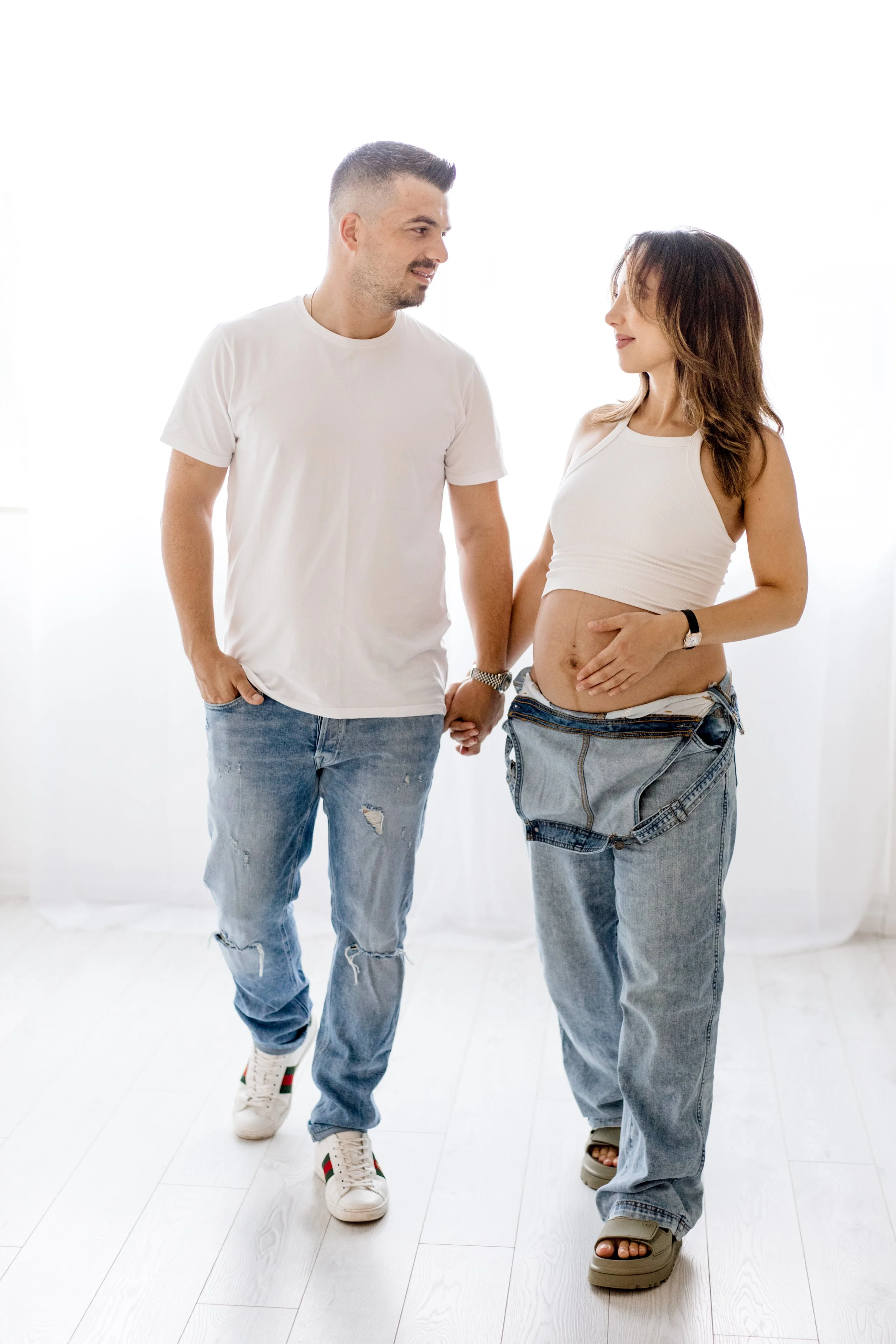 A pregnant woman and a man holding hands, walking together indoors with natural lighting.