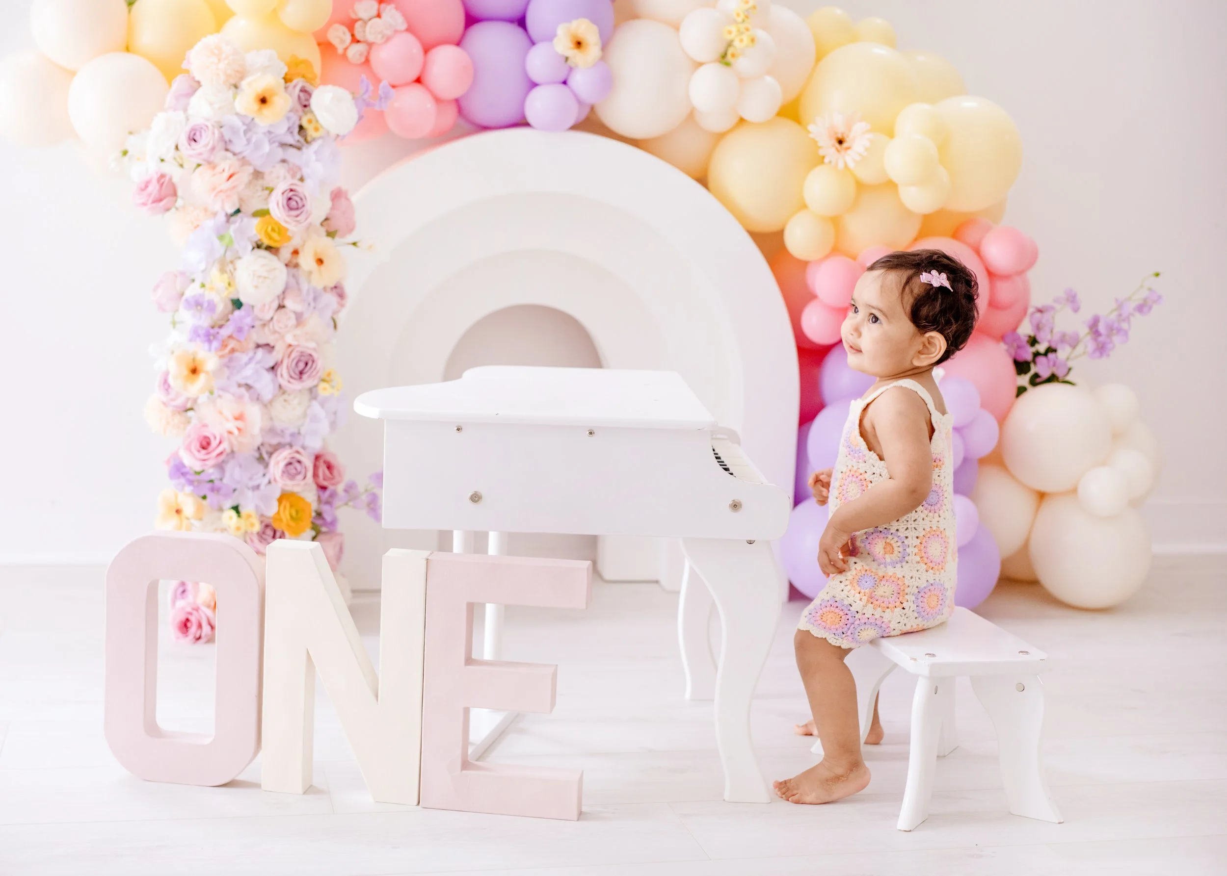 A little girl with short curly hair sit on a white bench next to a small white piano. Behind her is a backdrop of  balloons and a large decorative rainbow, Large block letters spell out "ONE" . first birthday photography Mississauga