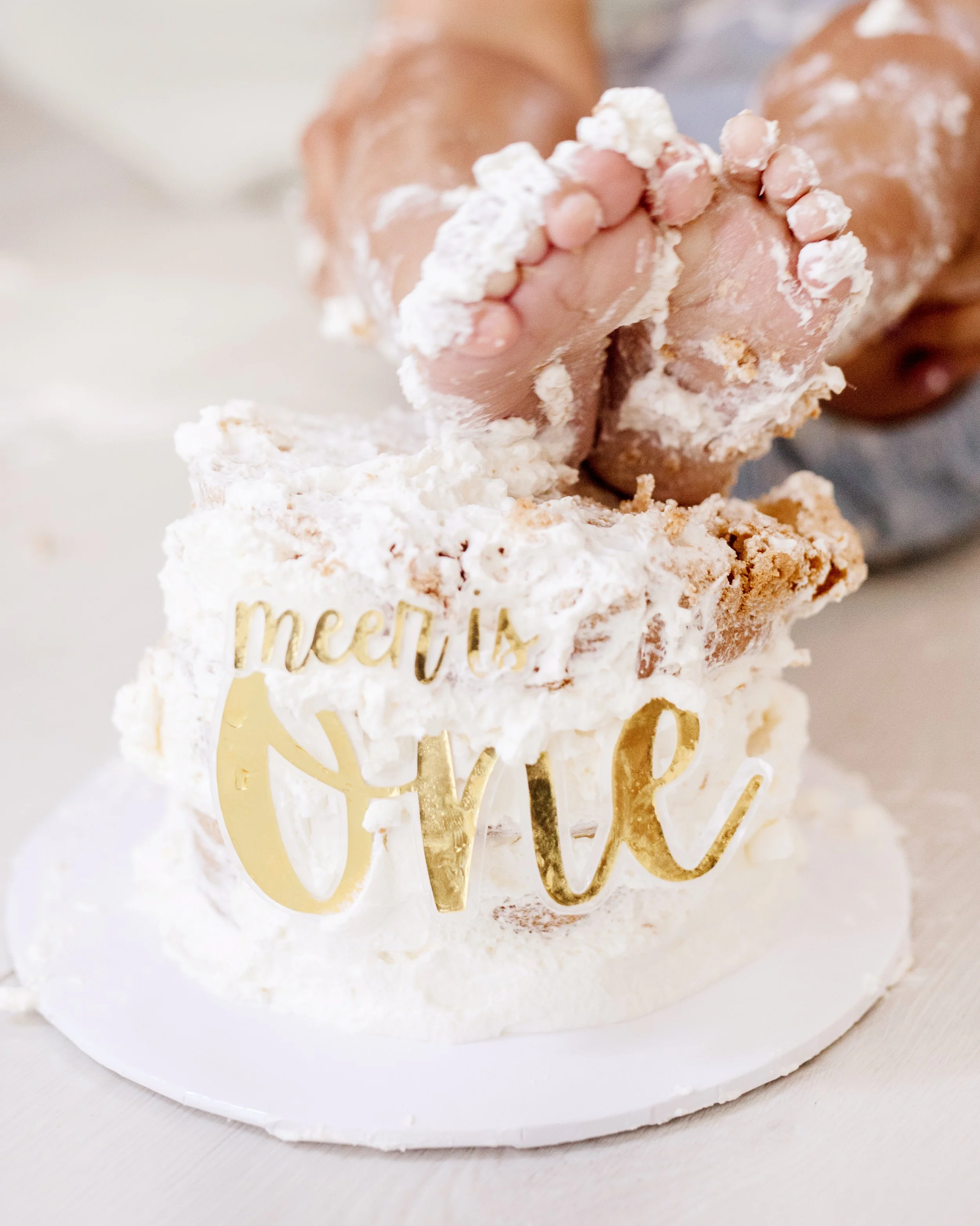 A small cake decorated with white frosting and a gold 'mom is loved' topper. Two baby feet covered in flour are positioned above the cake, suggesting a celebration of motherhood or a baby shower. .  first birthday cake smash Mississauga