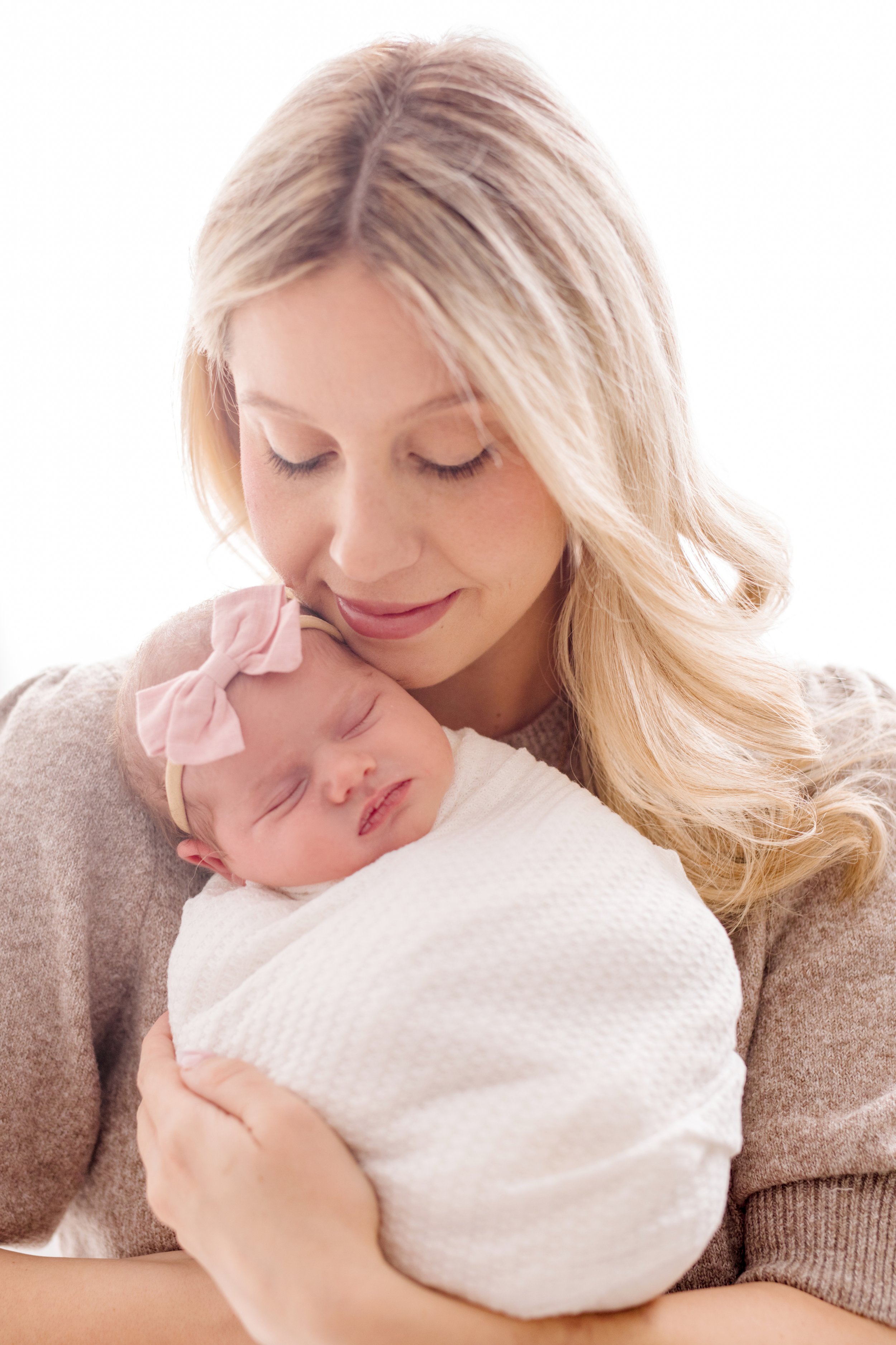 A woman with blonde hair holding a sleeping baby wrapped in a white blanket, with the baby wearing a pink bow headband.