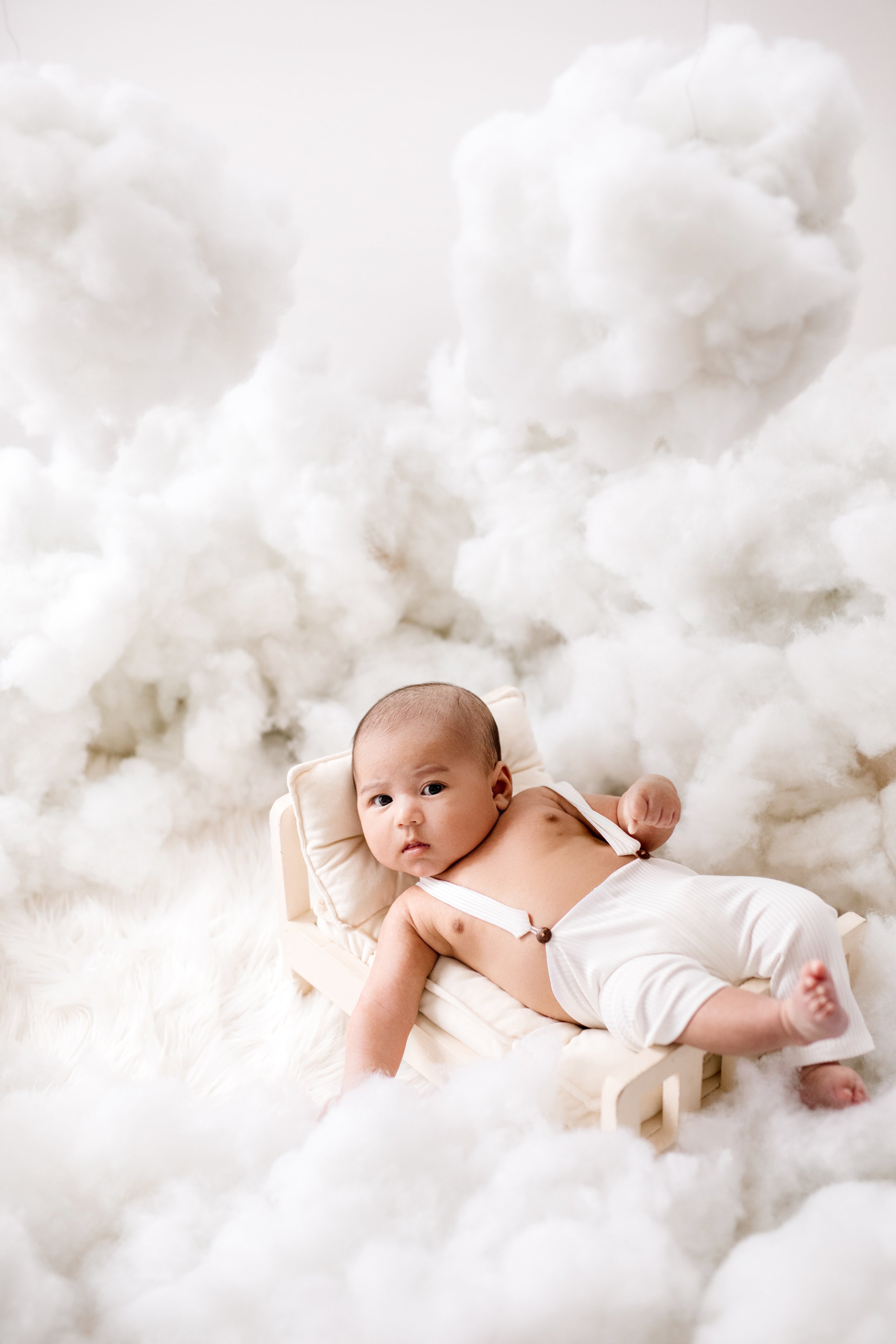 A baby lying on a small bed surrounded by fluffy white clouds, looking at the camera. professional baby photography in Mississauga.