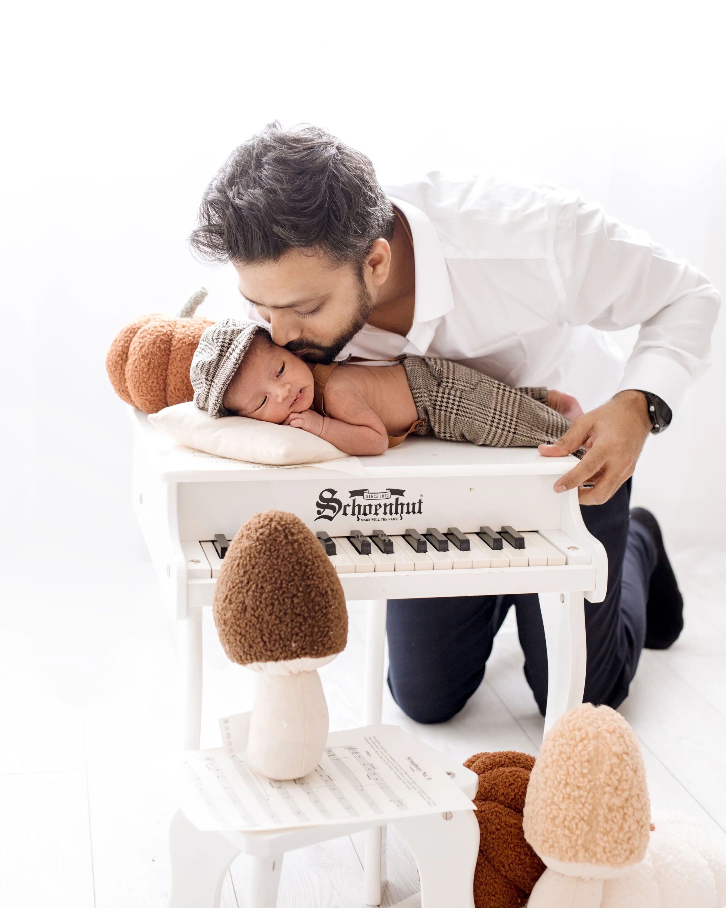 A father kisses a baby lying on a small white toy piano, surrounded by mushroom-shaped plush toys.