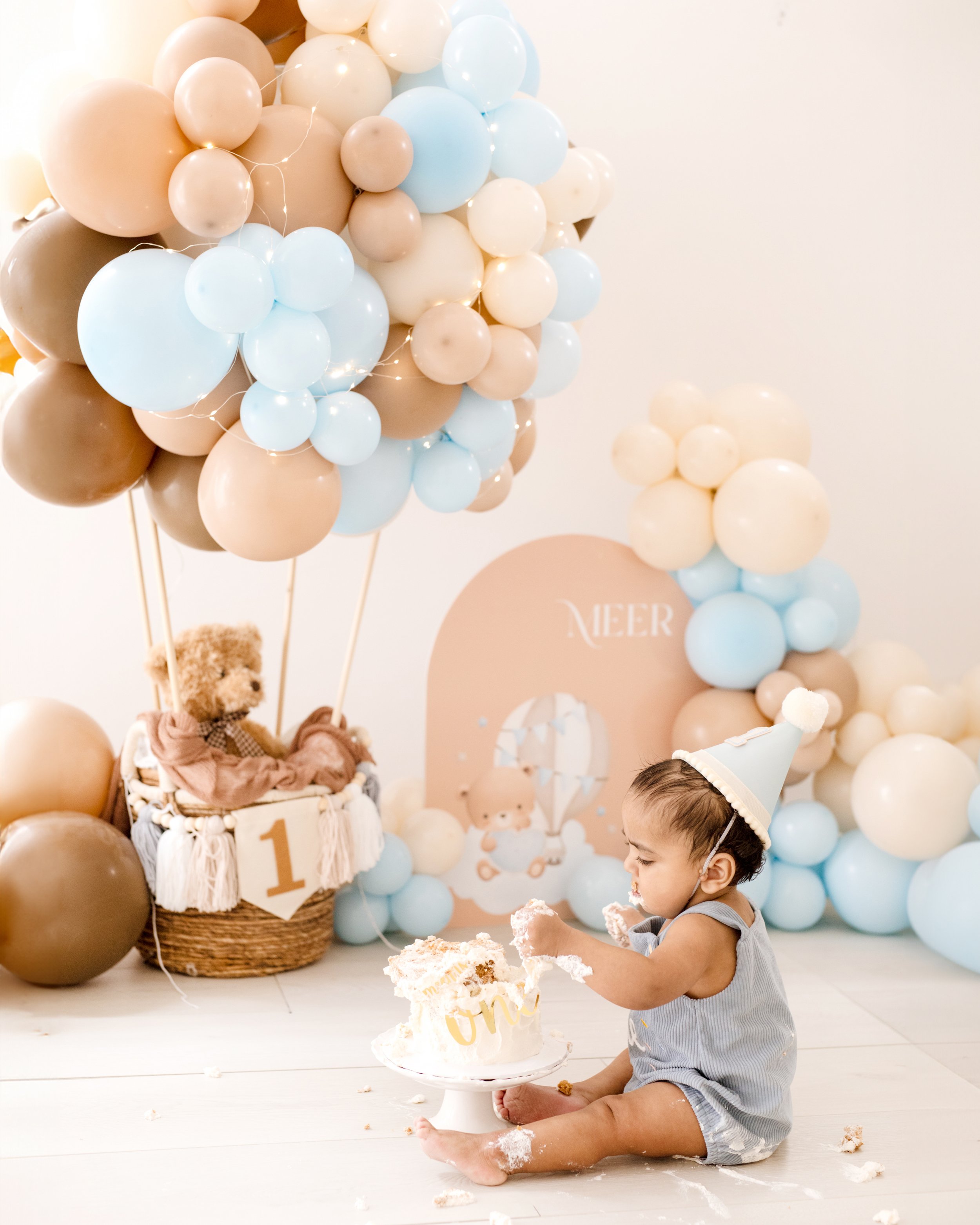 Young child wearing a party hat, sitting on the floor celebrating a first birthday with a cake, in front of balloon decorations and a backdrop with a hot air balloon and teddy bear theme. professional cake smash photography in Mississauga.
