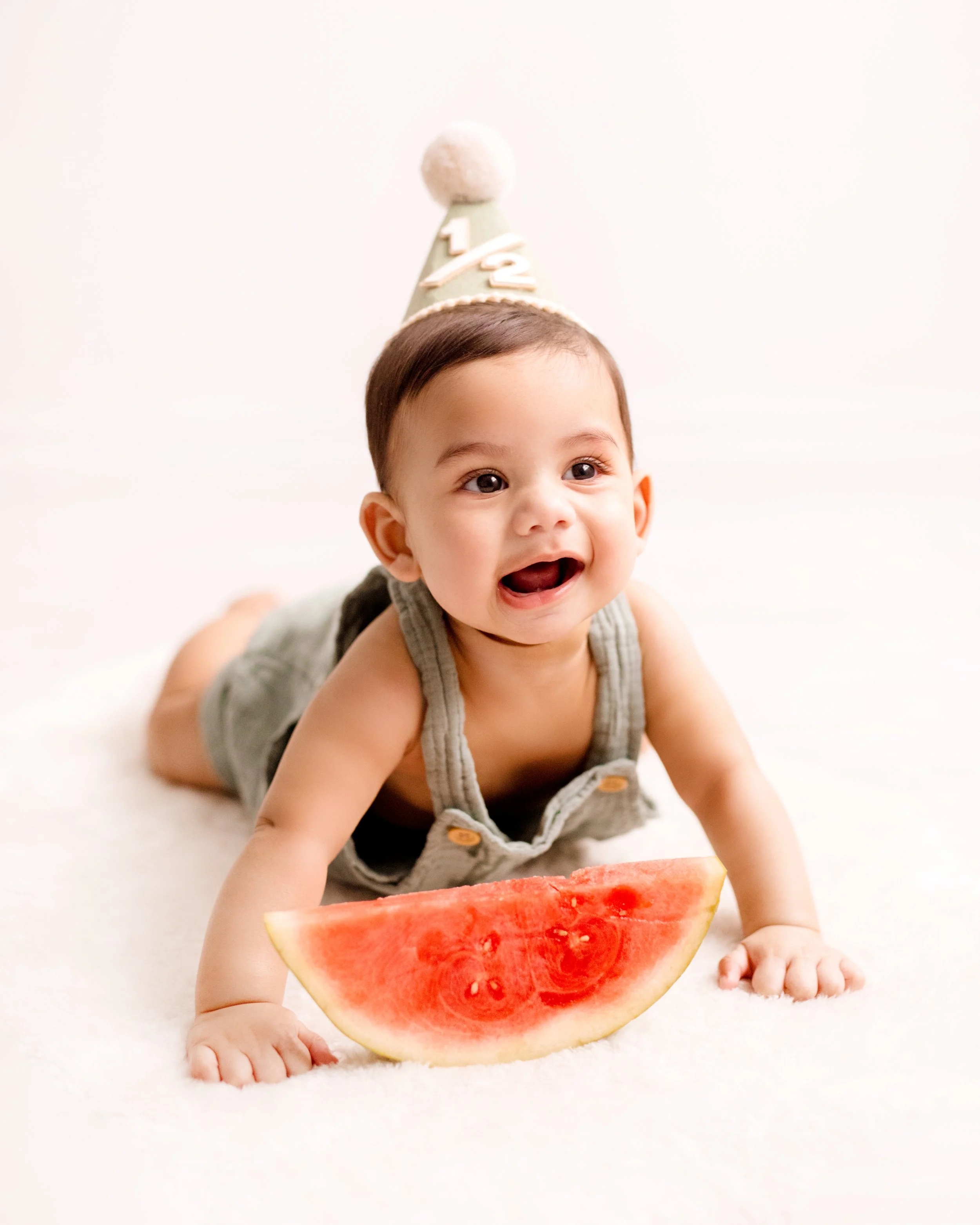 Six-month-old baby crawling on a white surface, wearing a party hat and holding a slice of watermelon – professional six month milestone baby photography in Mississauga.