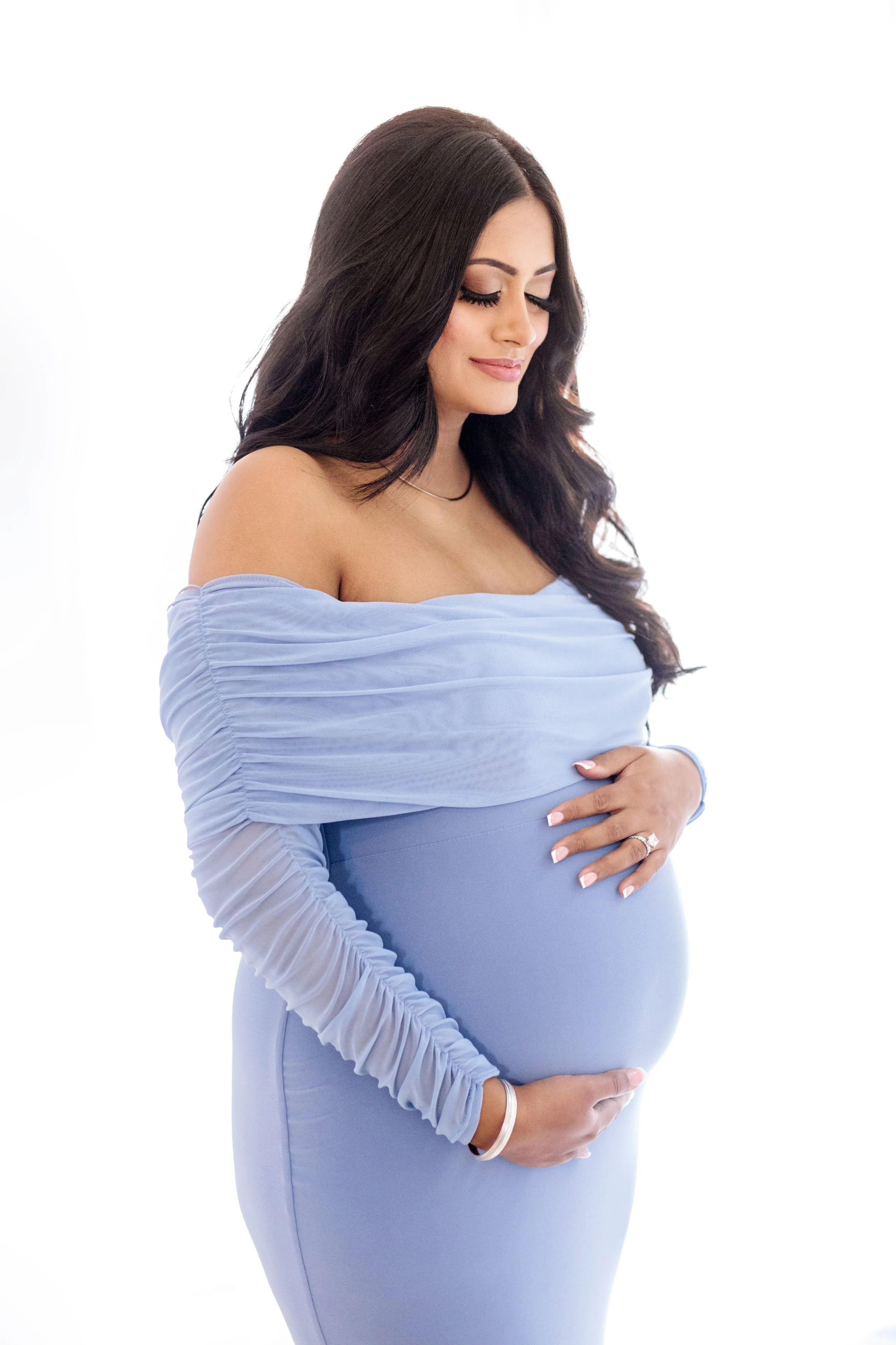 Pregnant woman with long dark hair in a blue off-shoulder dress gently holding her belly and smiling, photographed on a white background – professional maternity photography in Mississauga.