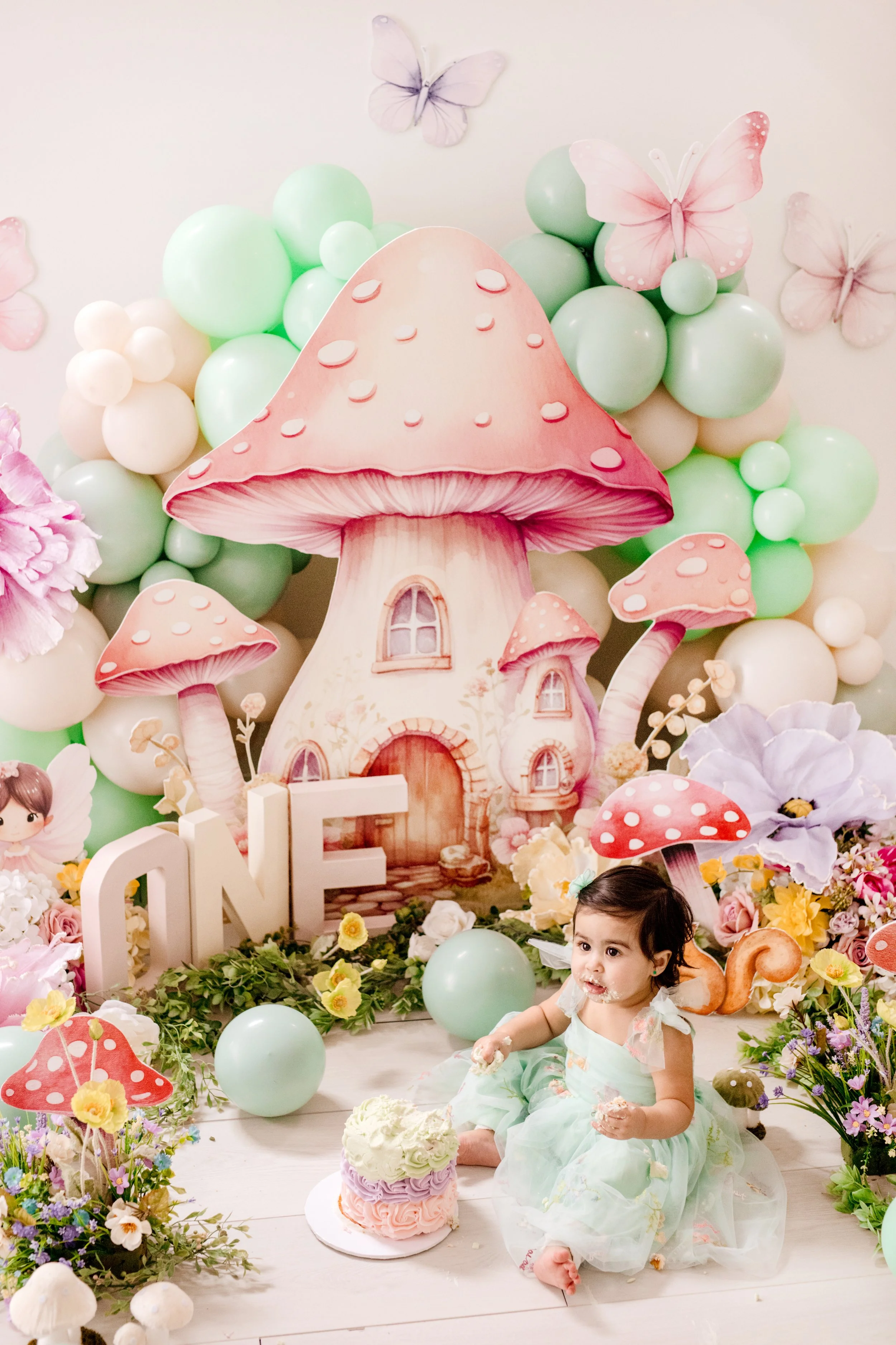 A girl in a mint green dress sitting on the floor at a birthday party with a coloured mushroom and fairy tale house backdrop, surrounded by flowers, balloons, and decorative mushrooms.  first birthday photographer Mississauga