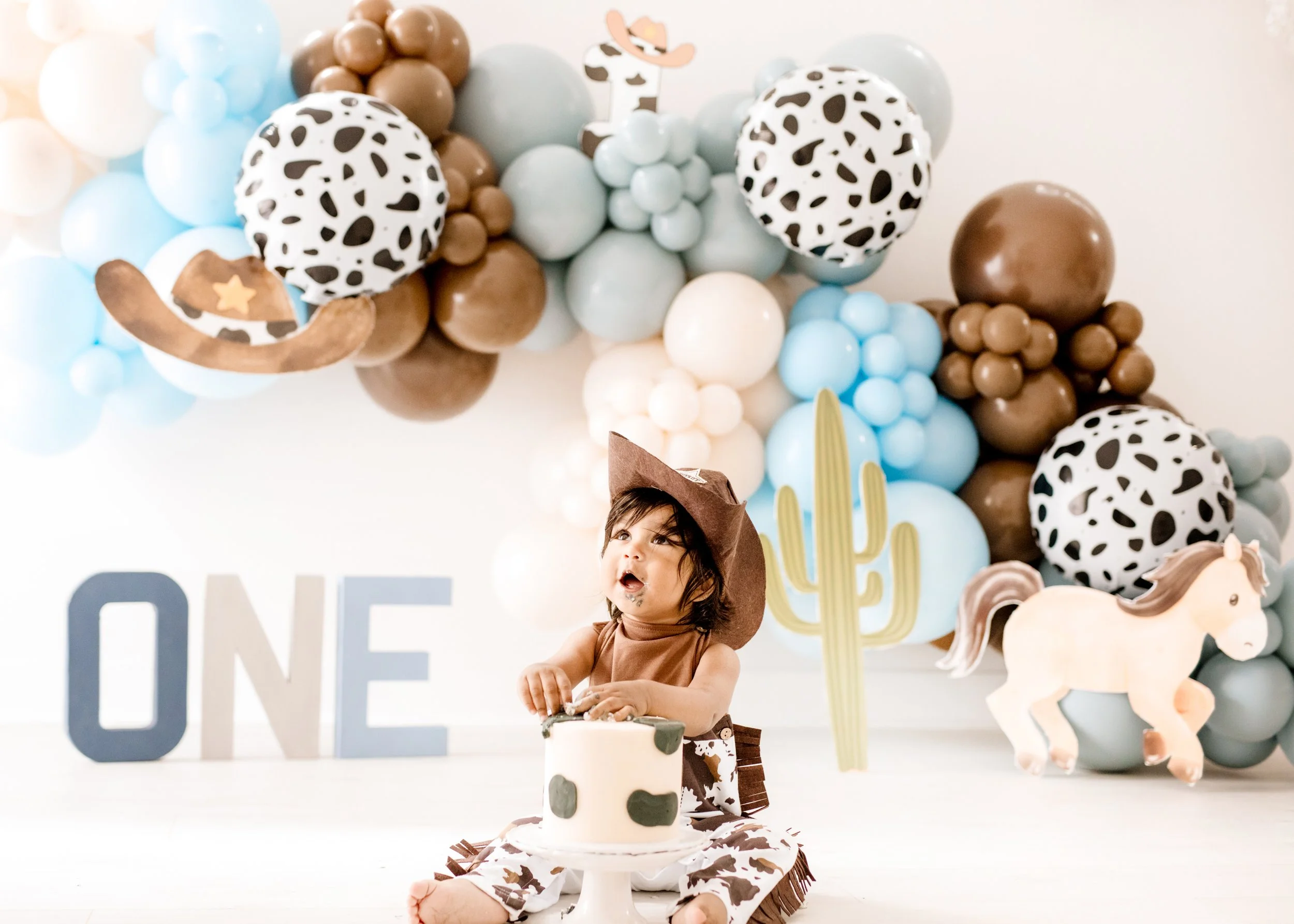 Young child celebrating a cow-themed birthday with a cake, cowboy hat, and farm decorations, including balloons with animal print, a cactus cutout, a horse decoration,  first birthday photoshoot Mississauga