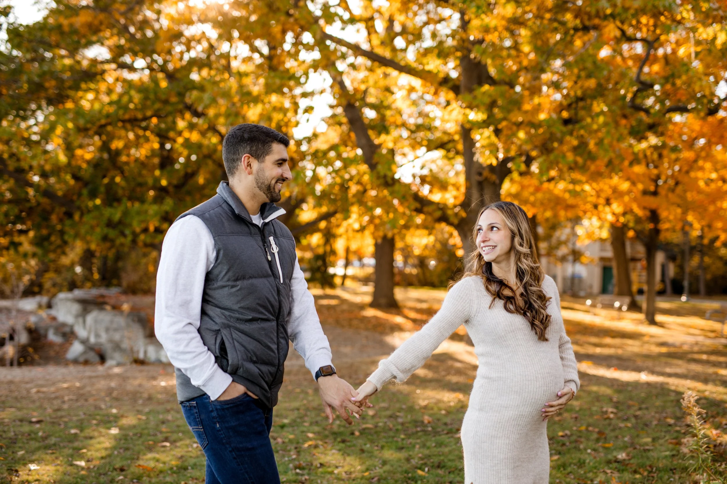 A man and a pregnant woman are holding hands and smiling at each other in a park with autumn trees in the background.