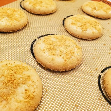 baked smores macaron shells piped on a silicone baking mat with graham cracker crumbs sprinkled on top