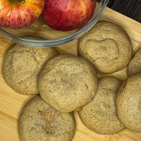 caramel apple cheesecake cookies on a wooden cutting board with a bowl of apples