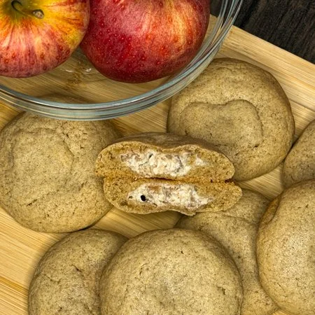 caramel apple cheesecake cookies on a wooden cutting board with a bowl of apples. one cookie is split in half so you can see the filling