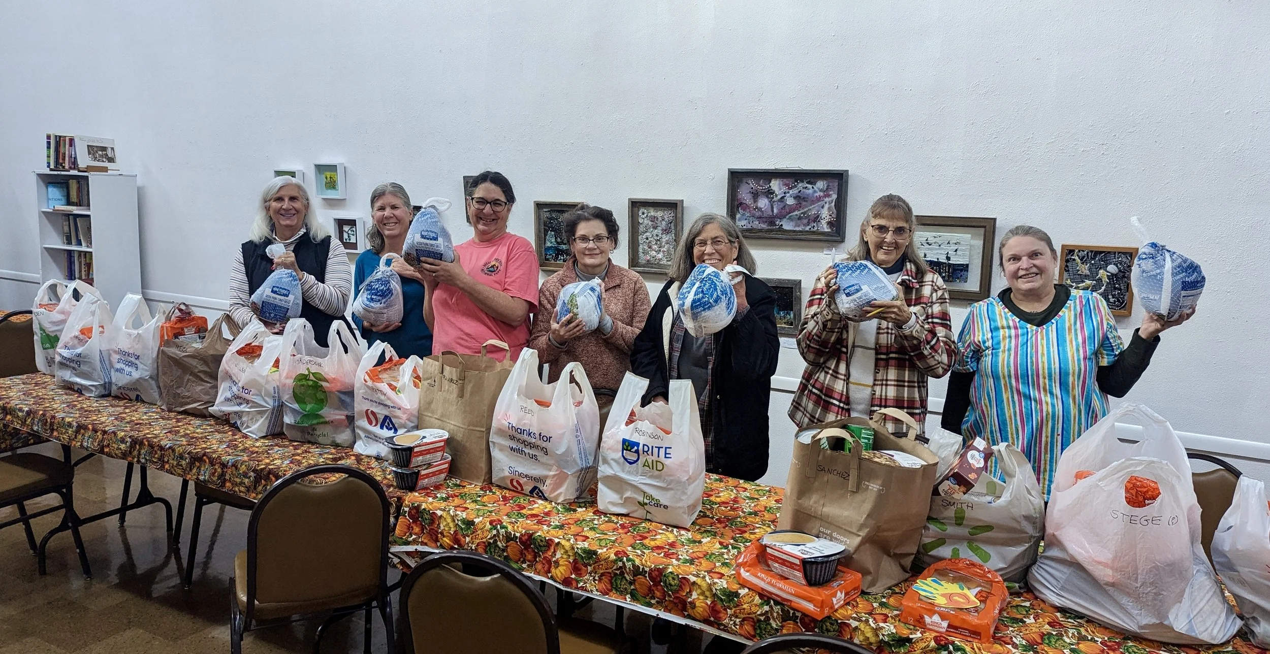 Food pantry volunteers (standing in front of art wall installation) surrounded by full grocery bags extend Thanksgiving turkeys toward the camera.