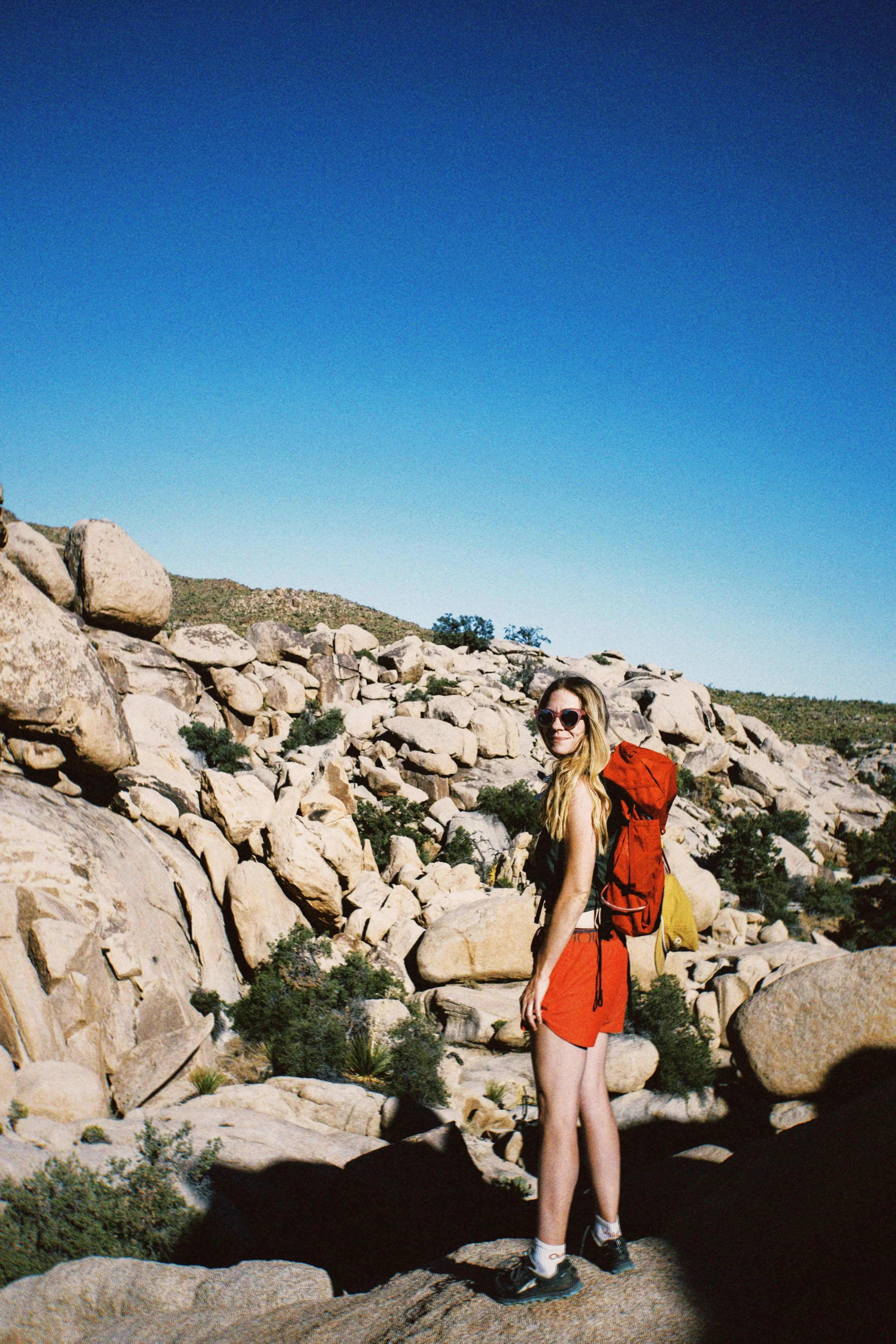 A young woman with long blonde hair, wearing sunglasses, a sleeveless black top, red shorts, and hiking shoes, stands outdoors among large rocks and desert vegetation under a clear blue sky.