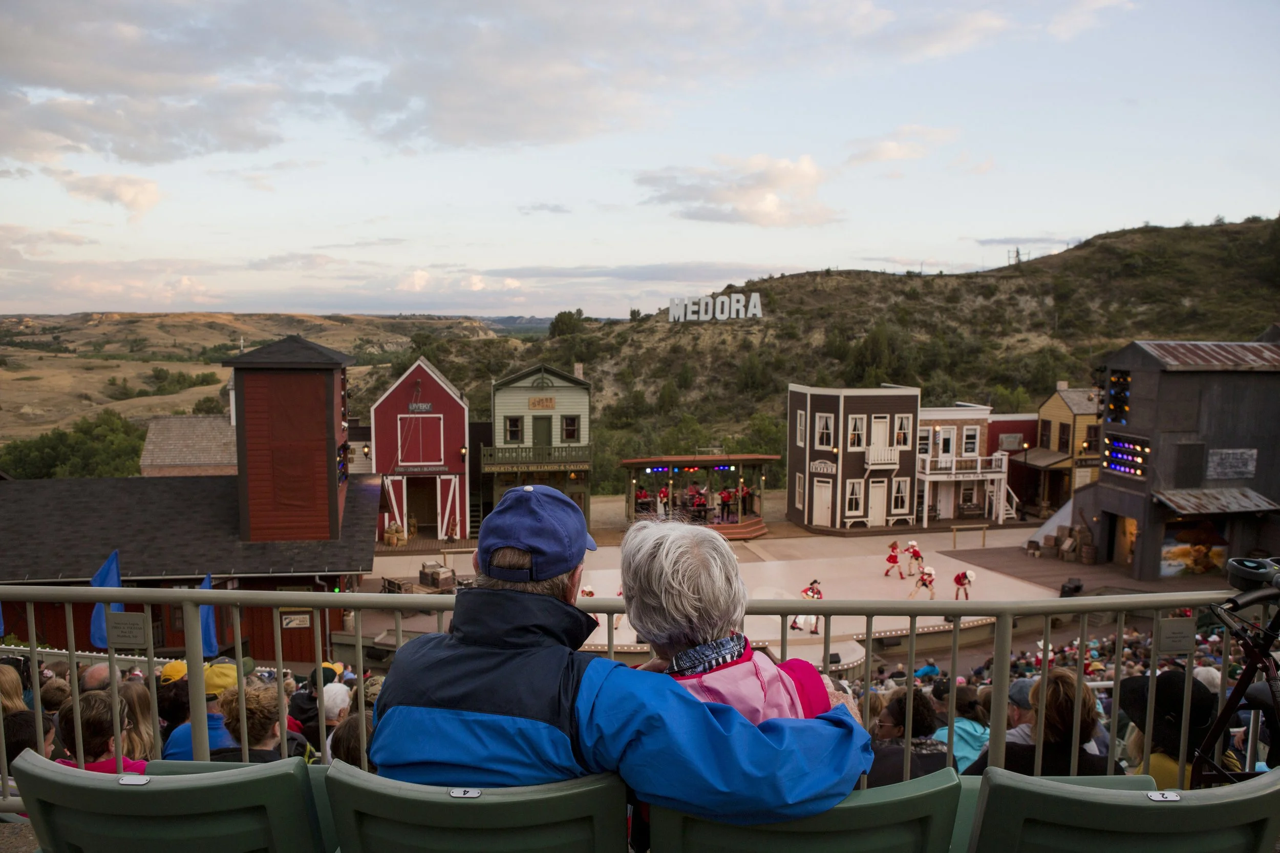 People seated in an outdoor amphitheater watching a stage performance set with western-themed buildings and a hillside with 'Medora' sign.