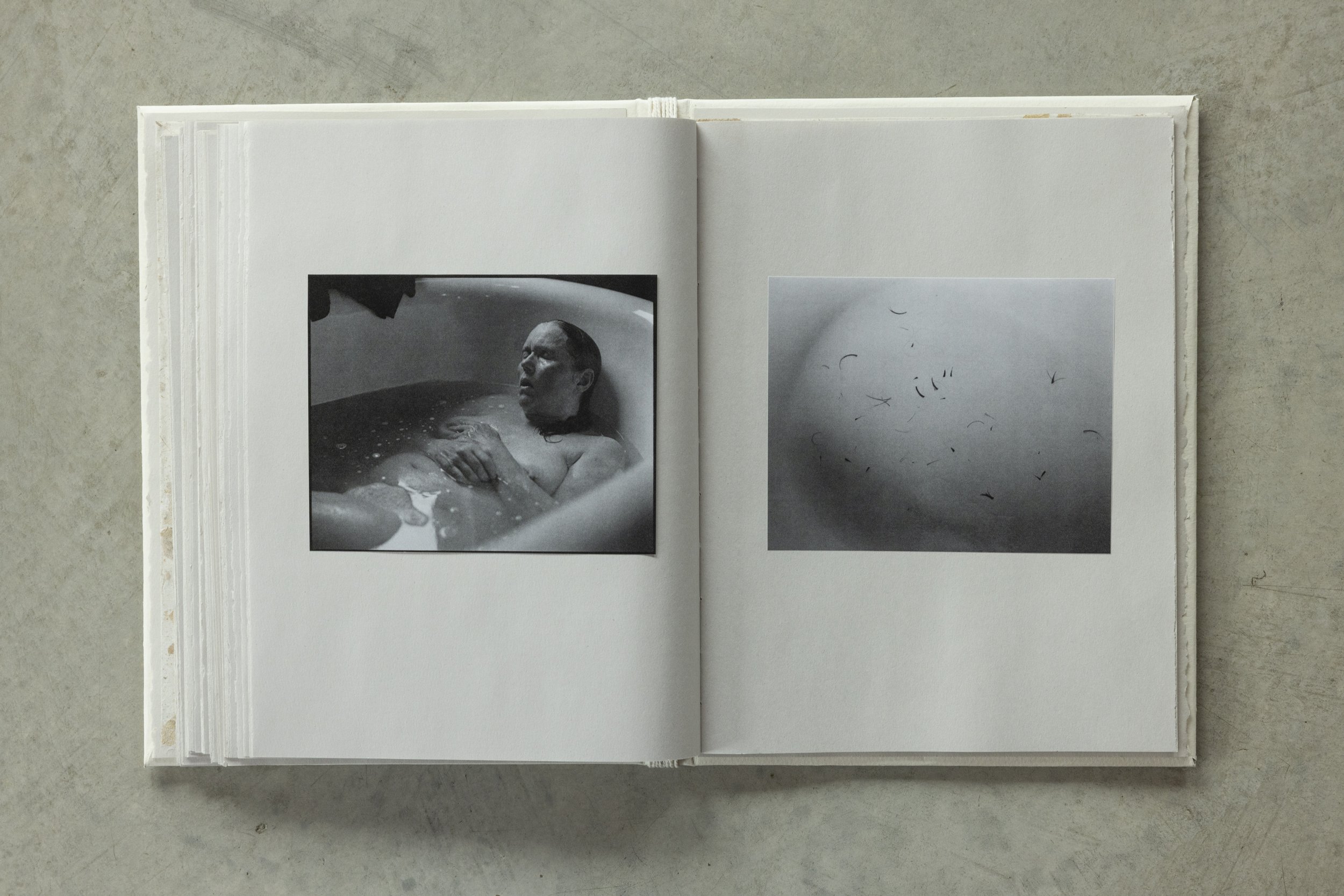 Open book on a table displaying black and white photographs of a woman in a bathtub and a close-up of feathers or hair.