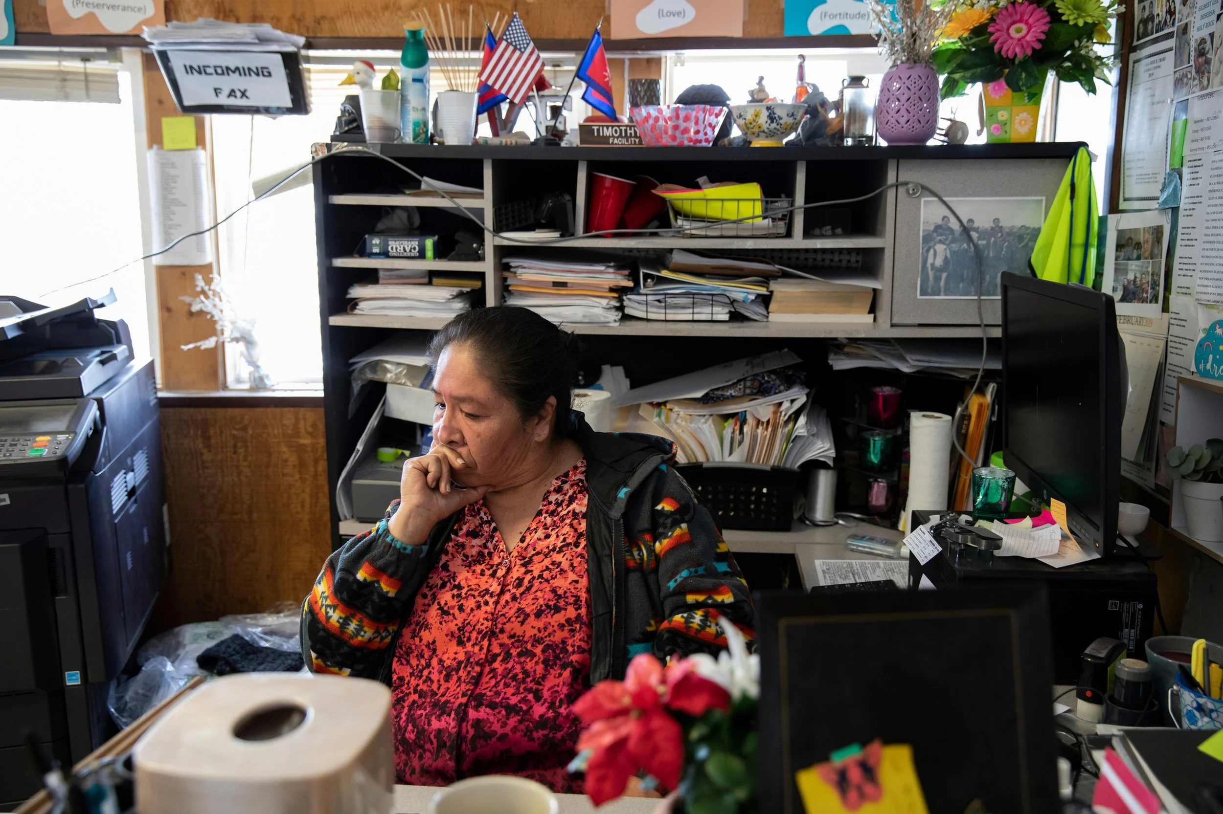 A woman with dark hair in a ponytail, wearing a colorful, patterned jacket and a red floral top, sitting at a cluttered desk in an office or workspace, with shelves filled with papers, folders, and office supplies behind her, focusing on her work.