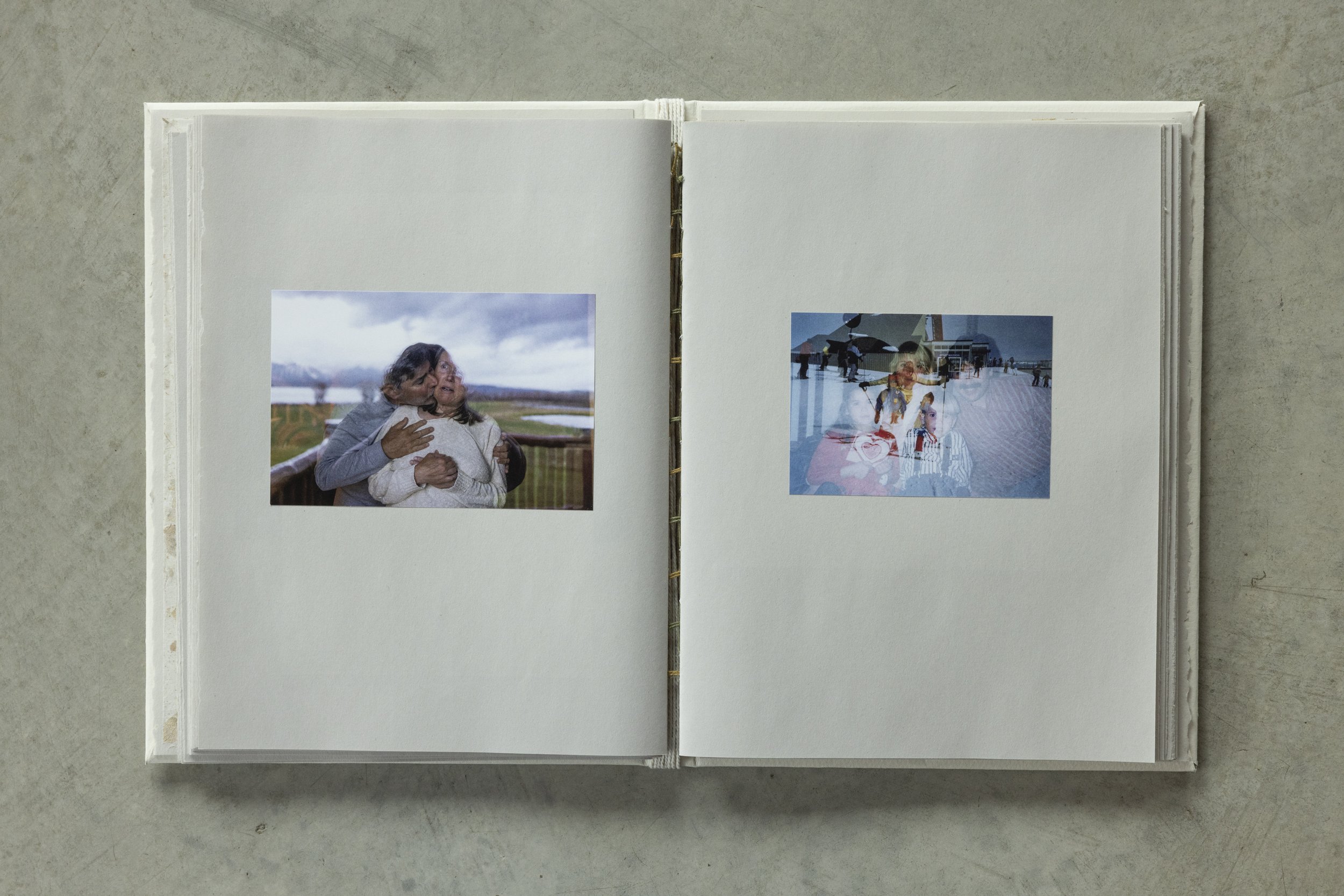 Open white photo album on a gray surface, showing two photographs: one of two people embracing outdoors with cloudy sky background, and another of people at an outdoor event with umbrellas.