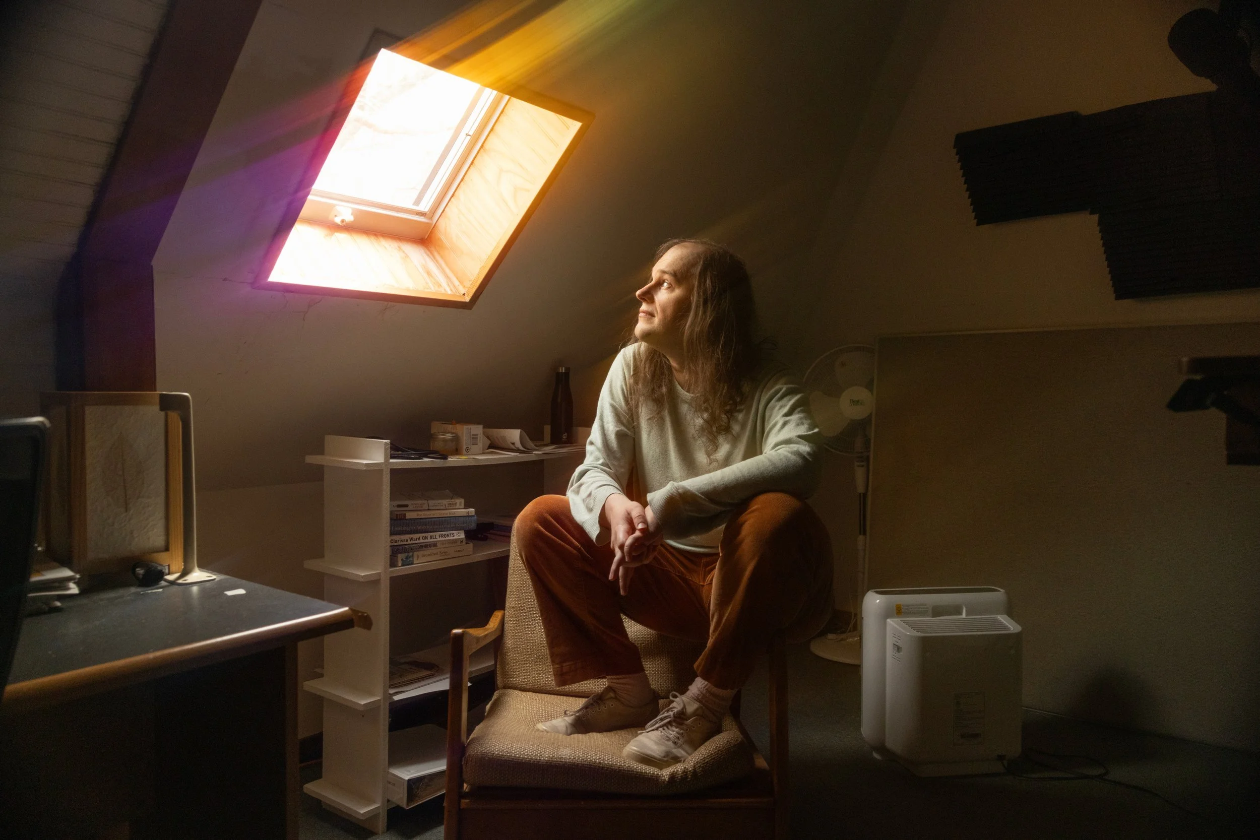 A woman with long brown hair, wearing a gray sweatshirt and orange pants, sitting on a chair with her legs crossed and feet resting on a cushioned stool, in an attic room with a sloped ceiling, illuminated by light coming through a skylight window in