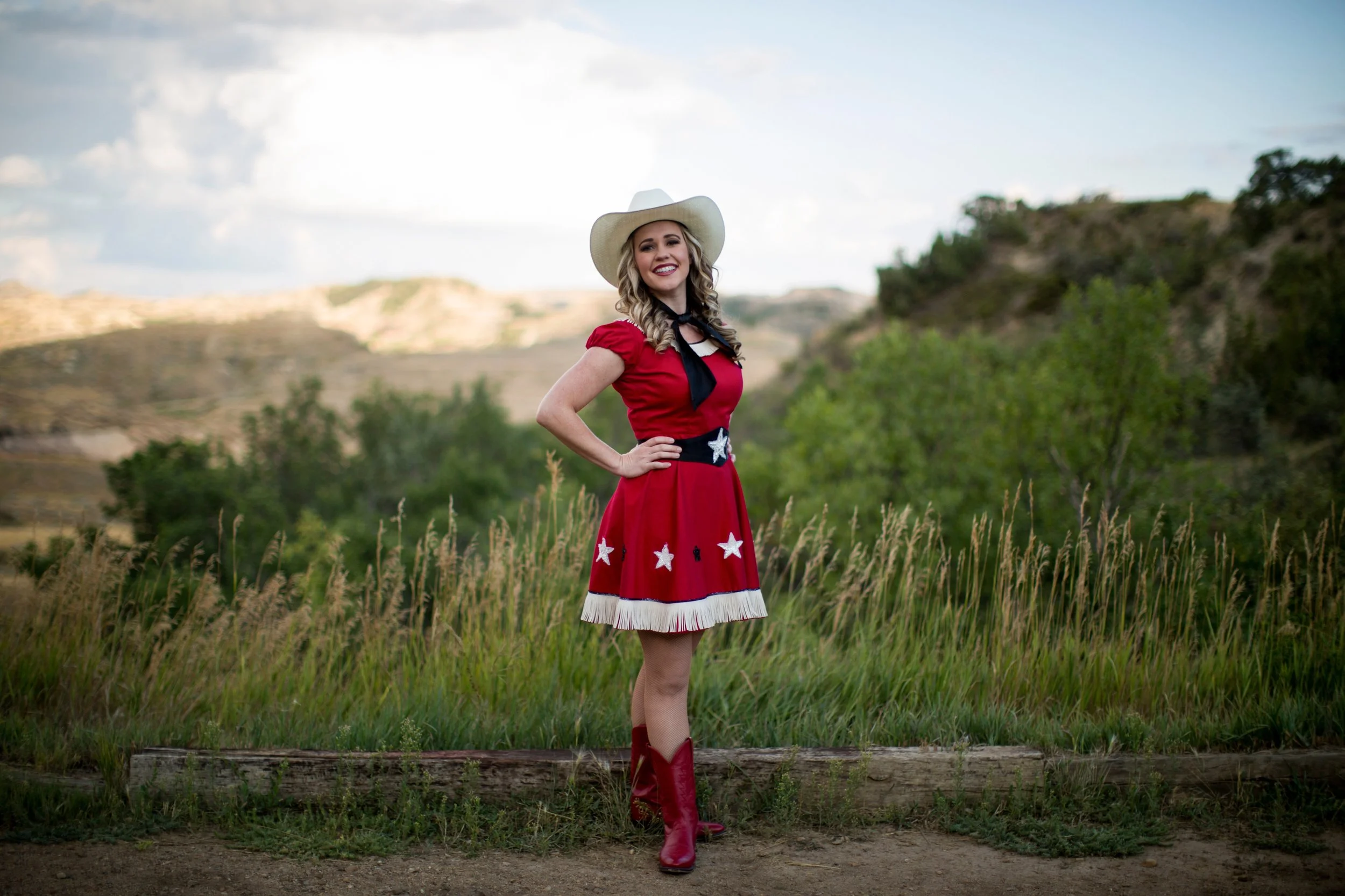 A woman standing in a grassy field wearing a red dress adorned with white stars, cowboy boots, and a large white cowboy hat, smiling with hand on hip, with rolling hills and cloudy sky behind her.