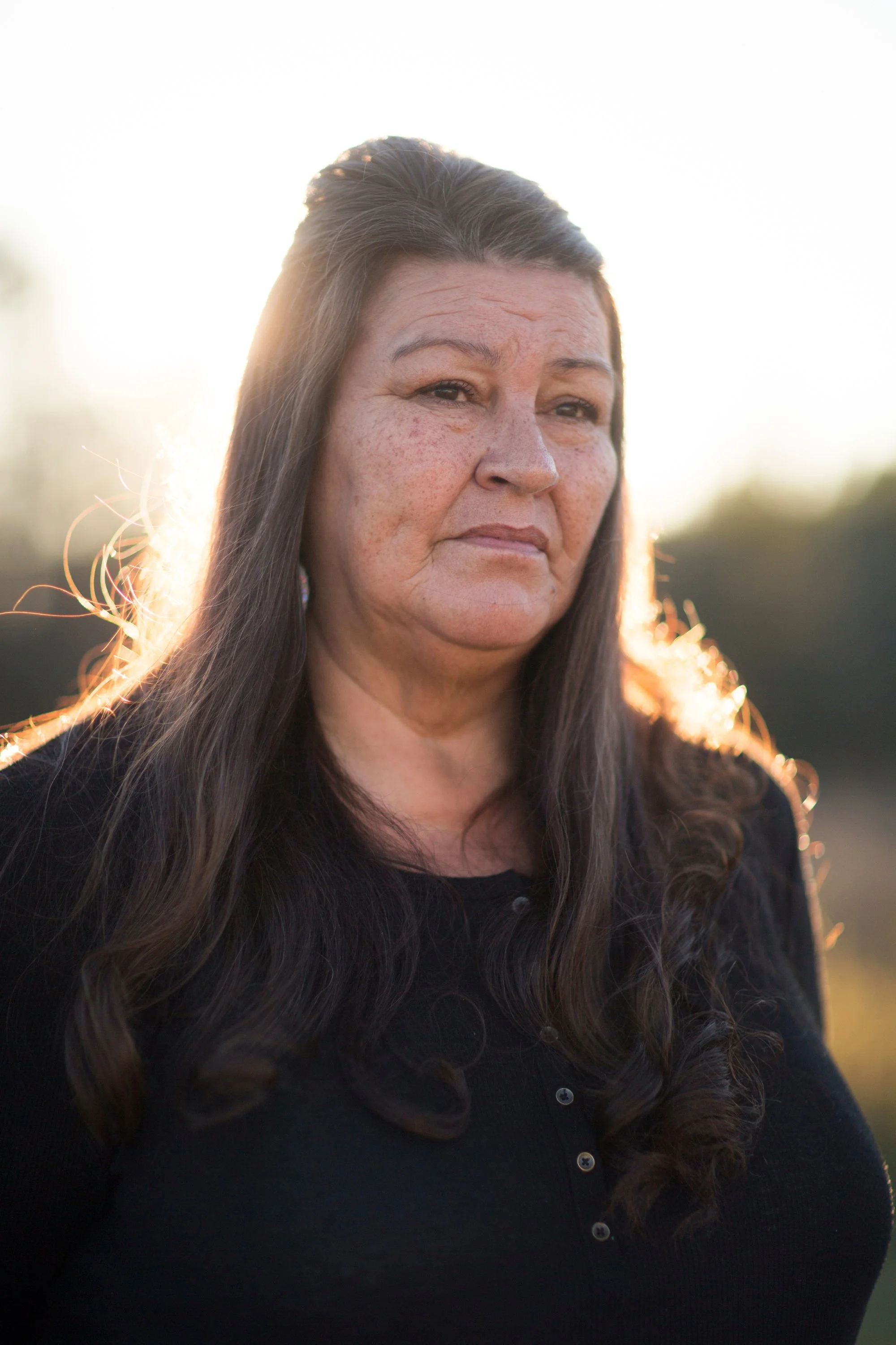 Close-up of a woman with long dark hair, wearing a black top, standing outdoors during sunset, with soft natural light illuminating her face and hair.
