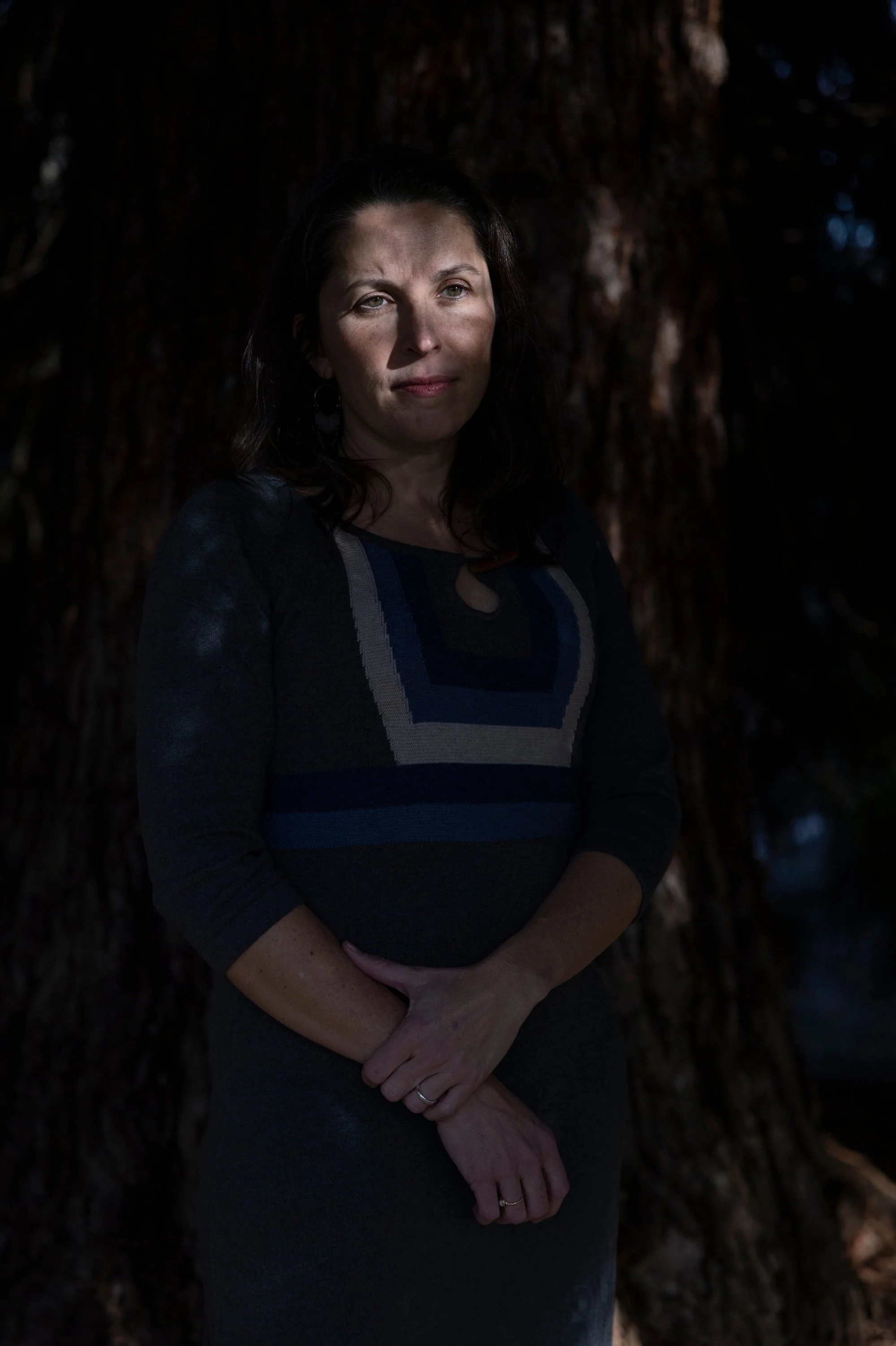 A woman with dark hair and light skin standing outdoors in front of a large tree, with sunlight partially illuminating her face.