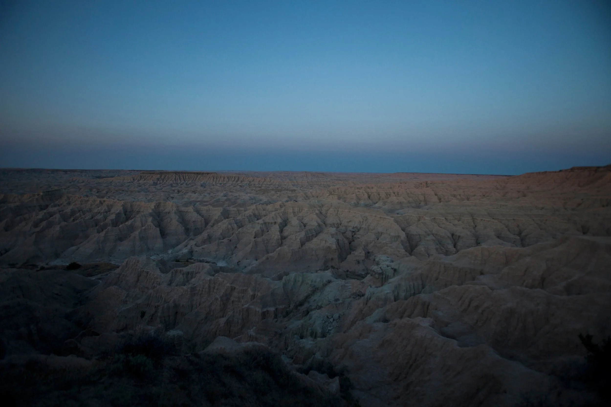View of a barren landscape with rugged, eroded hills under a clear blue sky during dusk or dawn.