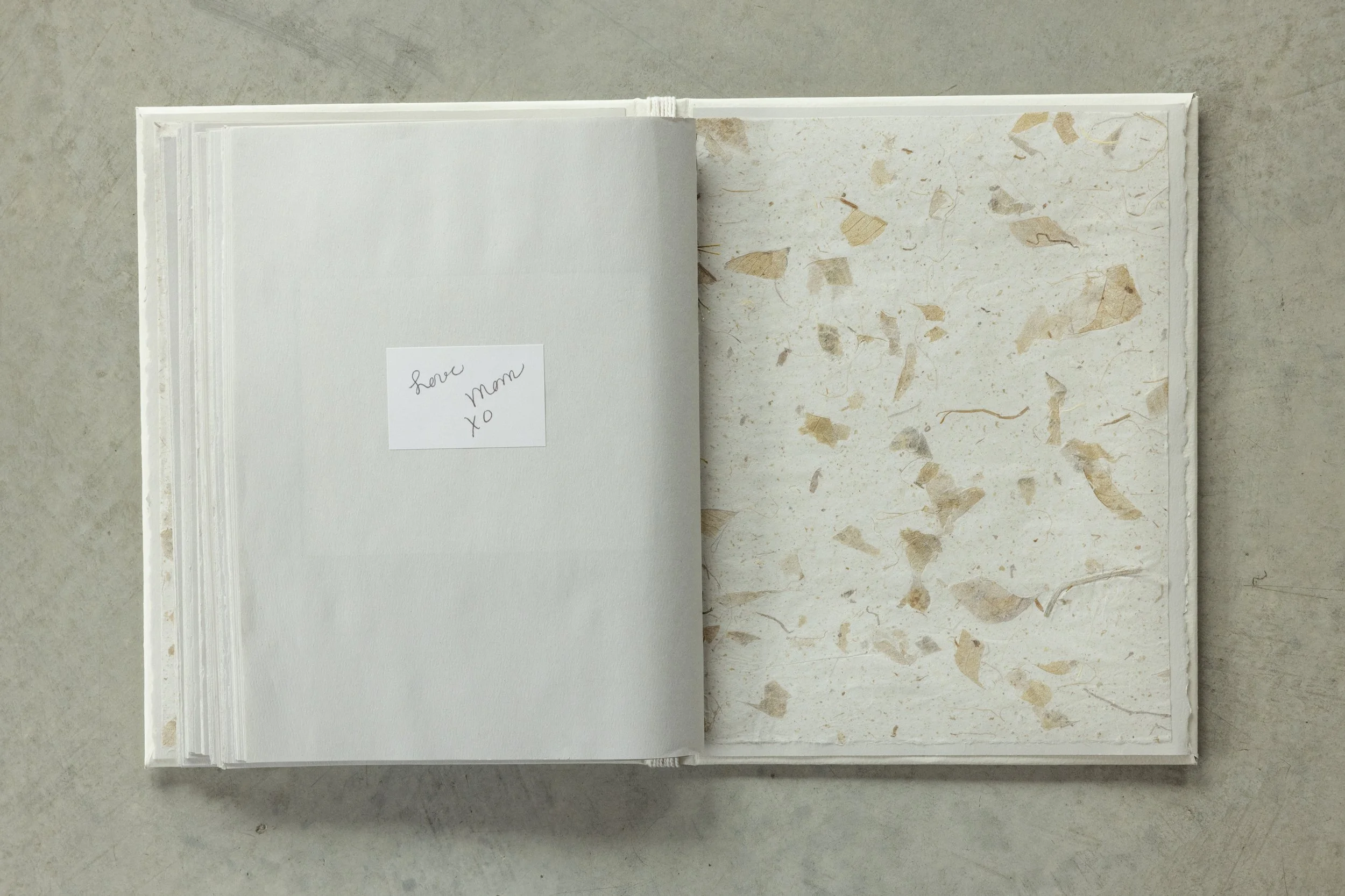 Open book with a handwritten note on the left page that says "love mom xoxo"; the right page features a marble pattern with beige and gray flecks on a light background, all on a gray surface.