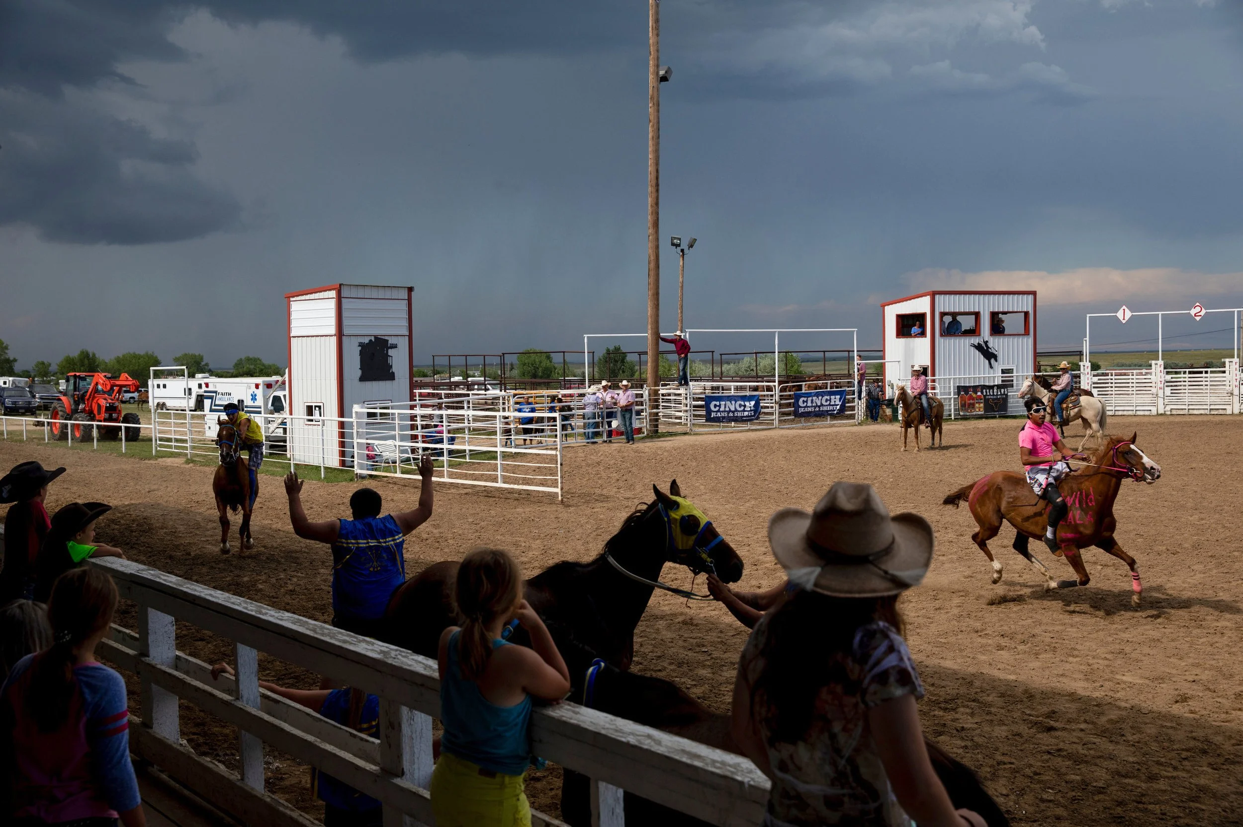 A racing event with people riding horses and watching from behind a white fence. Dark clouds are overhead, and there are concession stands and sponsors' banners in the background.