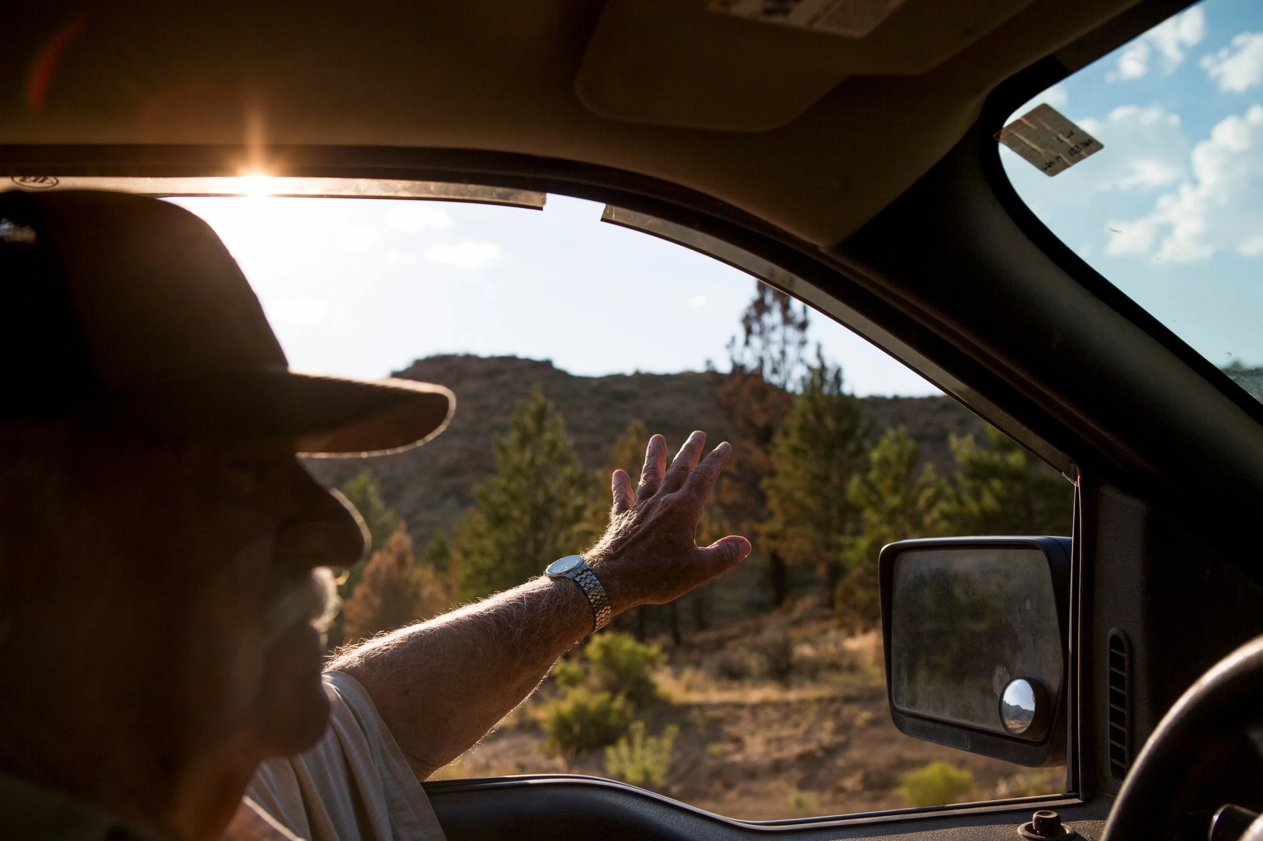 View from inside a vehicle with a person reaching out the window and wearing a hat and watch, overlooking a landscape with trees and a hill during sunset.