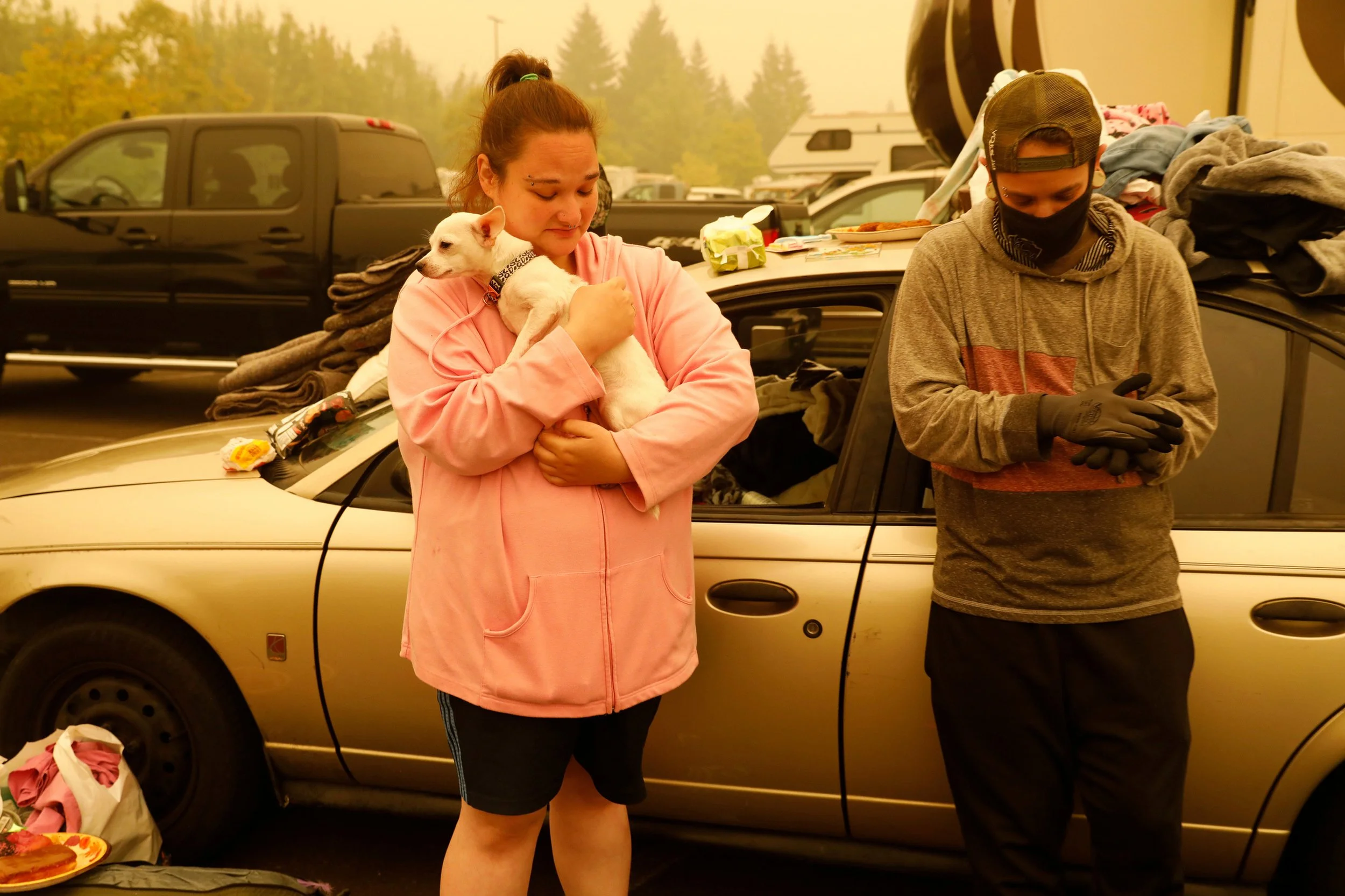 A woman in a pink hoodie holding a small white dog with a black collar, standing next to a man wearing a gray hoodie, black face mask, and a baseball cap, in a parking lot with cars and trees in the background.