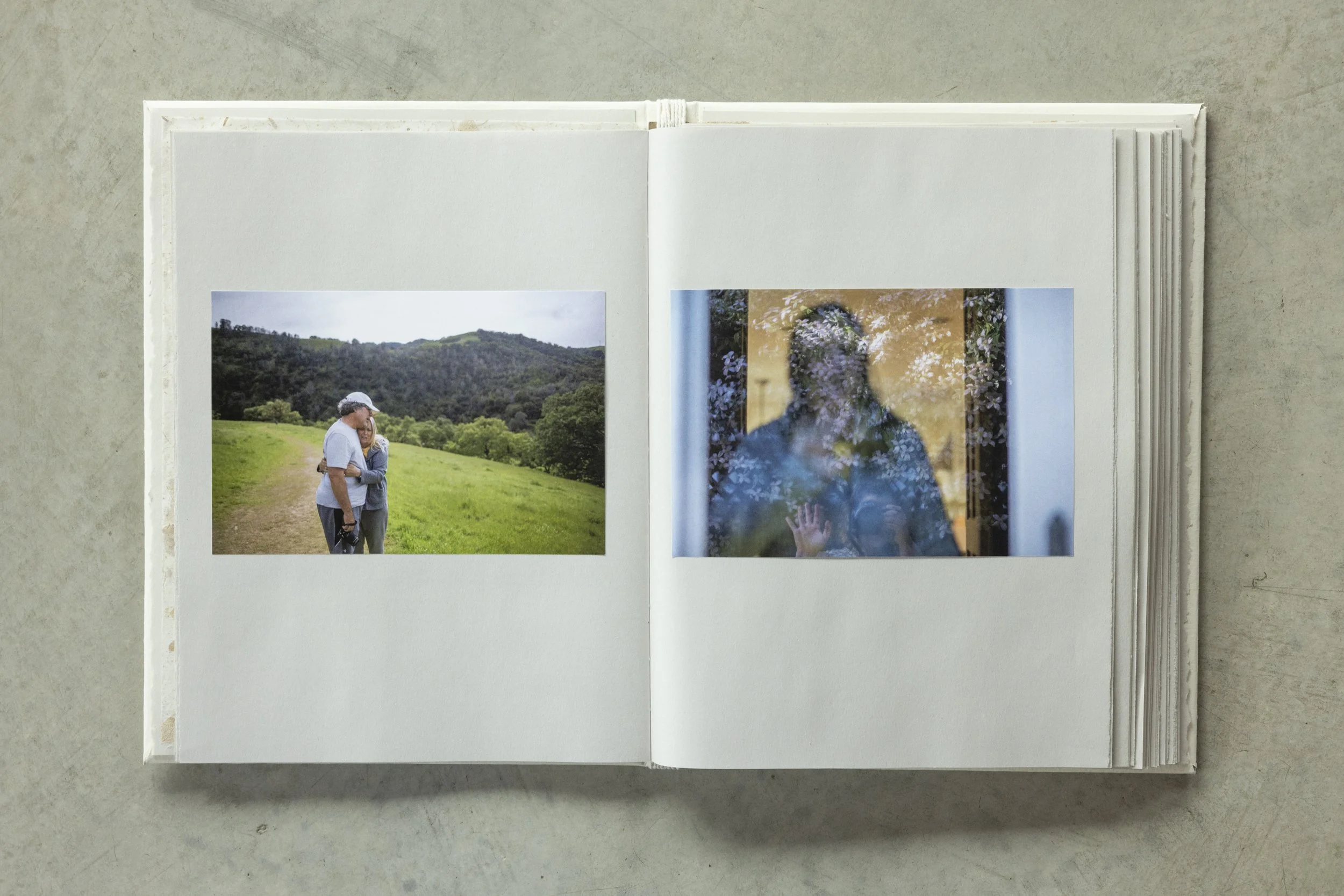 Open photo album showing two pages with photographs: one of a couple hugging outdoors in a green field and one of a woman behind a glass door with trees reflected on it.